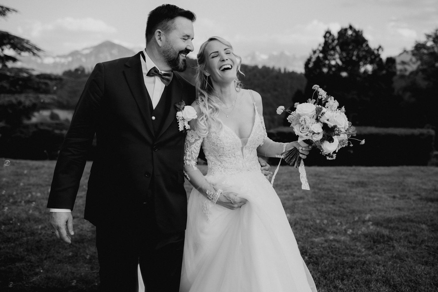 Black and white photo of laughing bride holding bouquet and groom in tuxedo on wedding day with mountains in background.