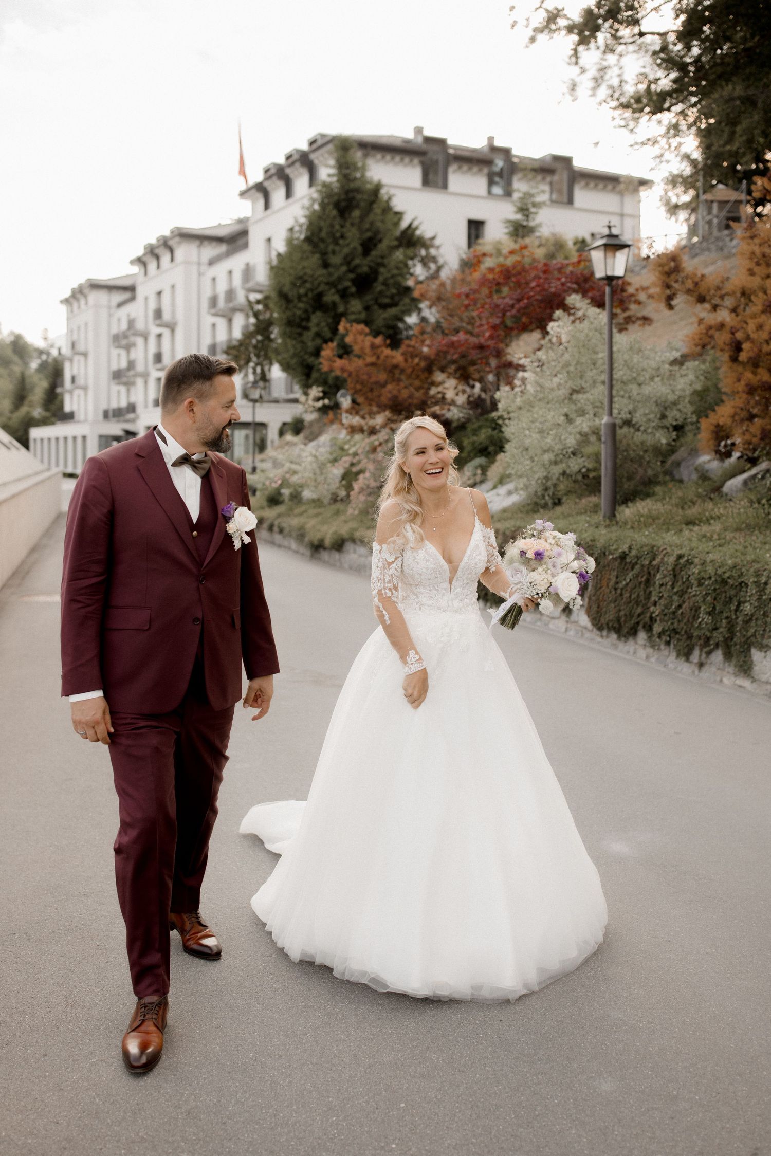 A couple in wedding attire walks along a path in front of a stately mountain hotel.