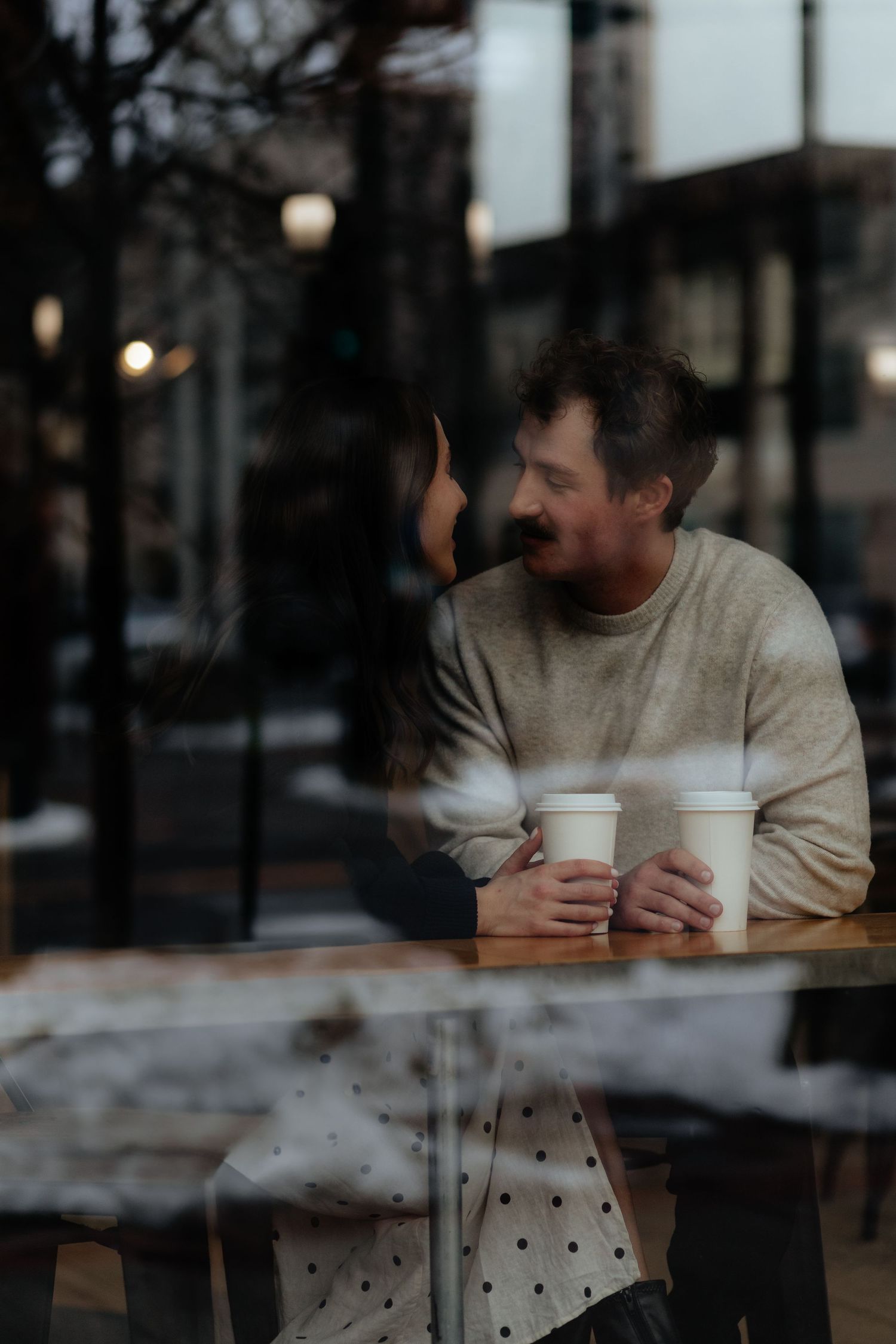 A couple shares a cozy moment over takeout coffee cups at a cafe window during dusk.