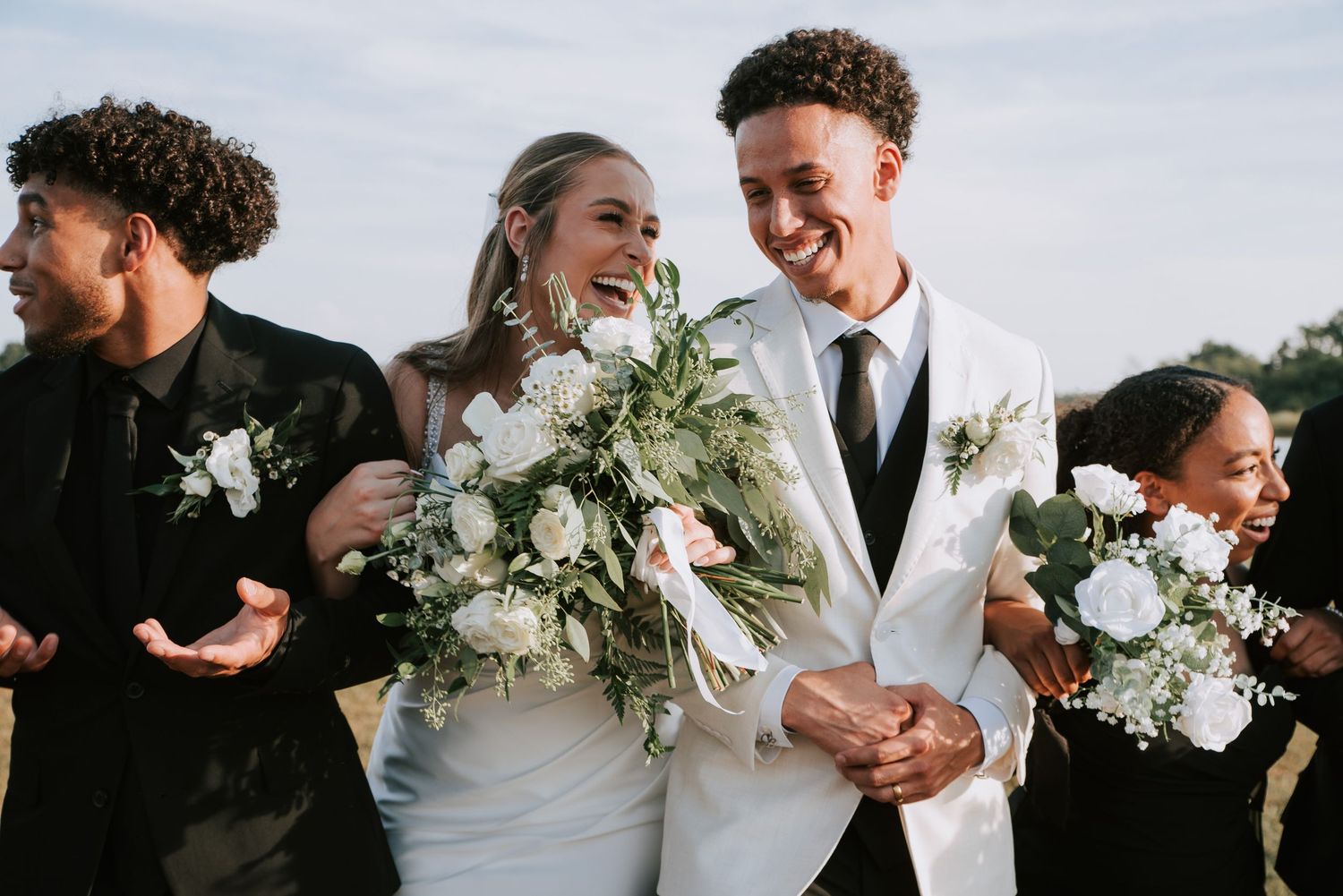 Happy interracial couple in wedding attire with white floral bouquets, celebrating with friends outdoors.