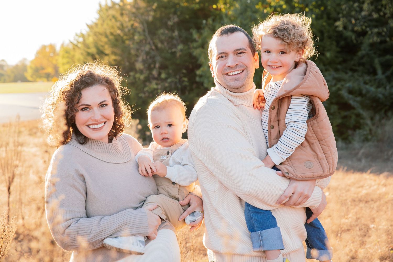 Happy family of four posing outdoors in golden sunlight, parents holding two young children in a scenic autumn field.