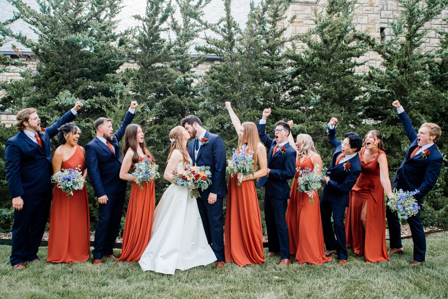 Wedding party in navy suits and rust orange dresses celebrating outdoors near evergreen trees.
