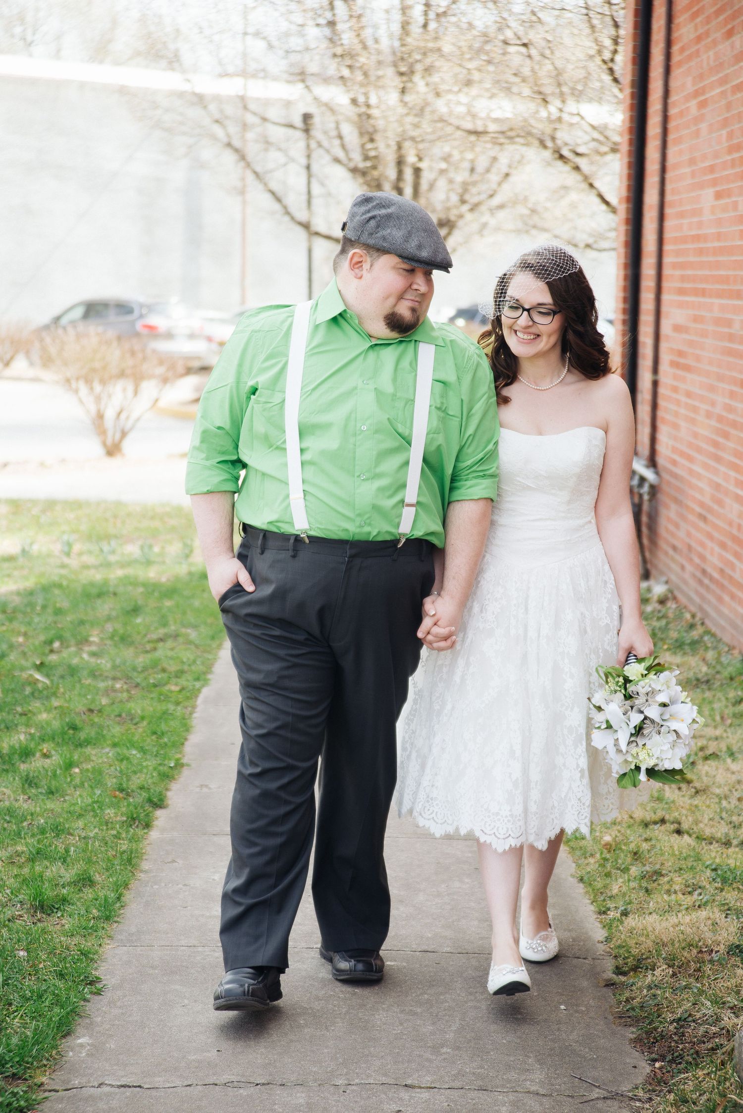 Newlywed couple walks hand-in-hand on a sidewalk, groom in green shirt with suspenders, bride in lace dress.