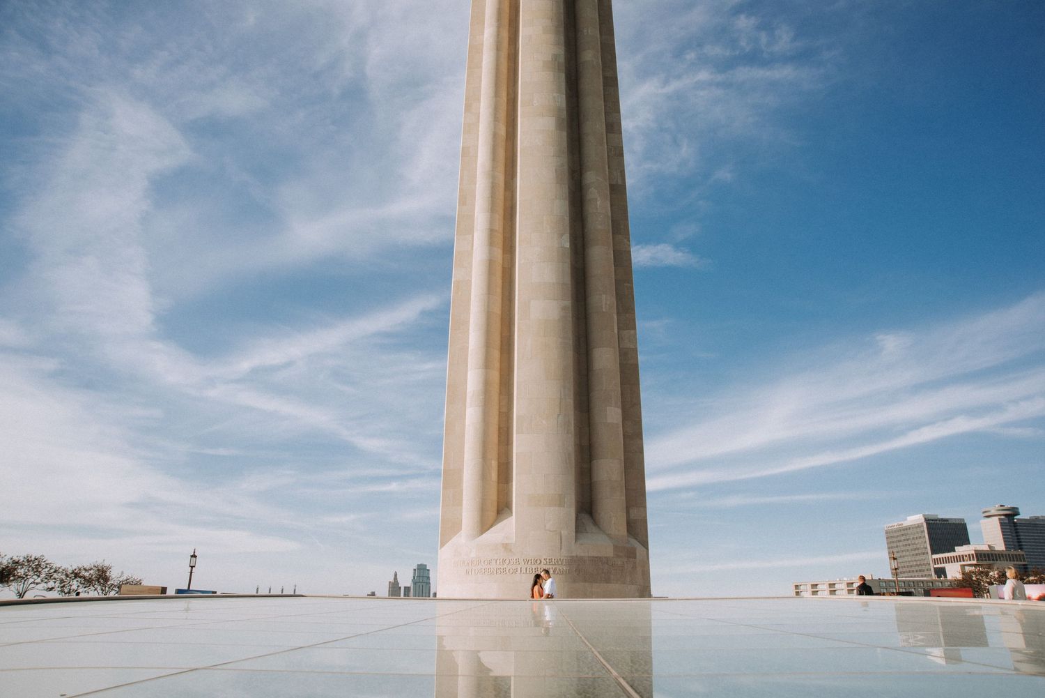 Couple sharing intimate moment beneath tall Liberty Memorial monument under blue cloudy sky.