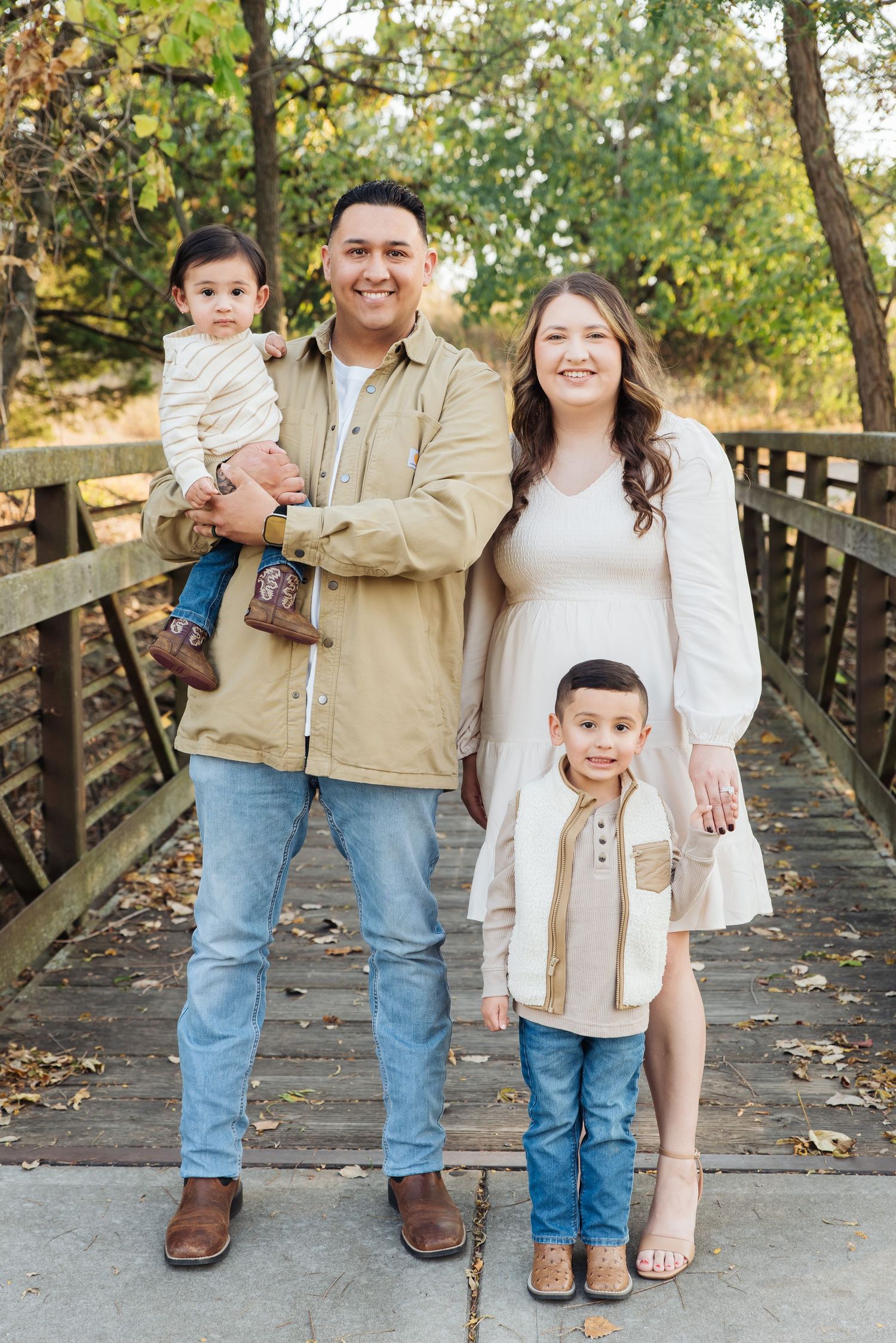 Happy family of four poses on a wooden bridge surrounded by autumn foliage in coordinating neutral outfits.