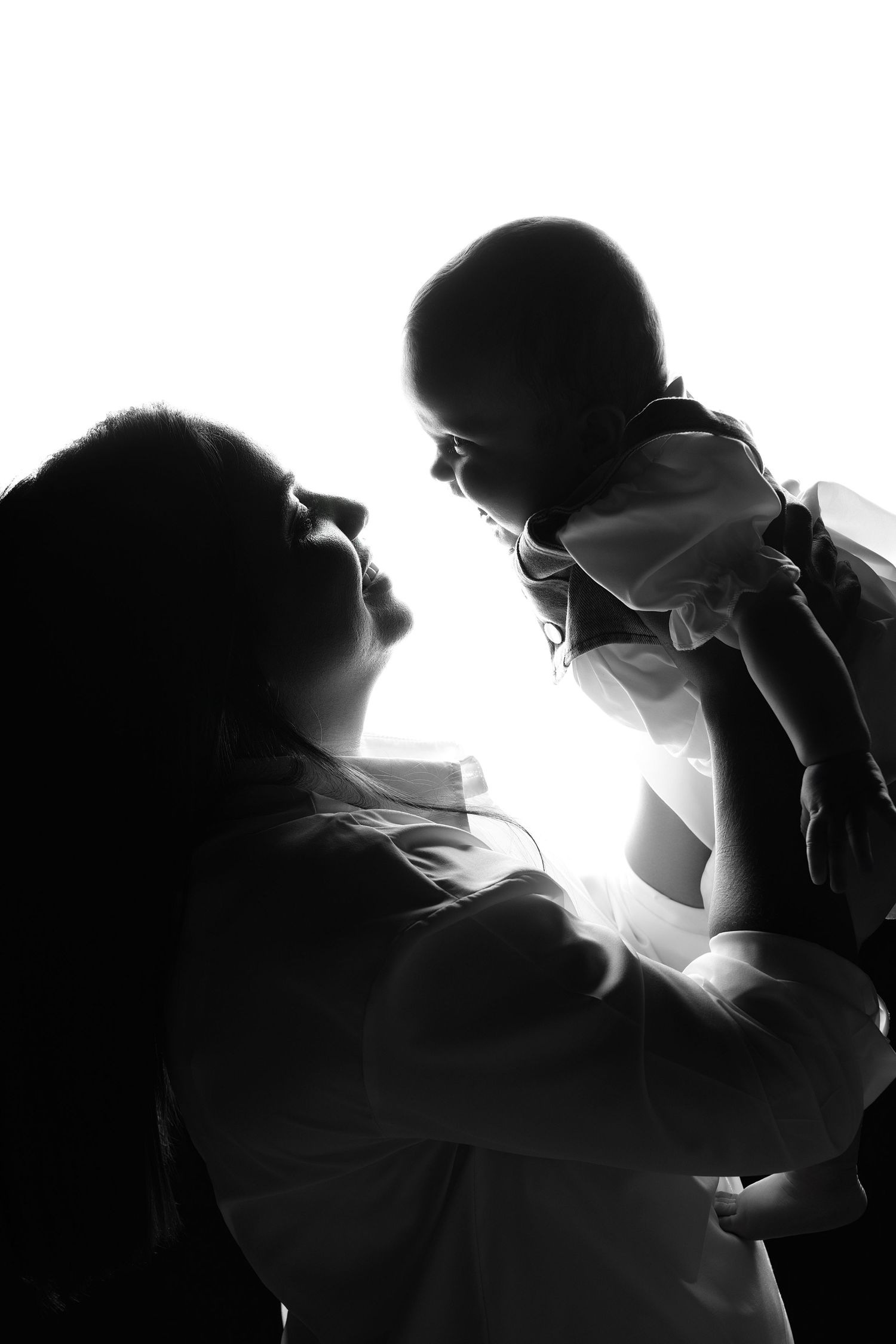 Black and white silhouette of a mother lifting her baby up in the air, backlit with bright white light.