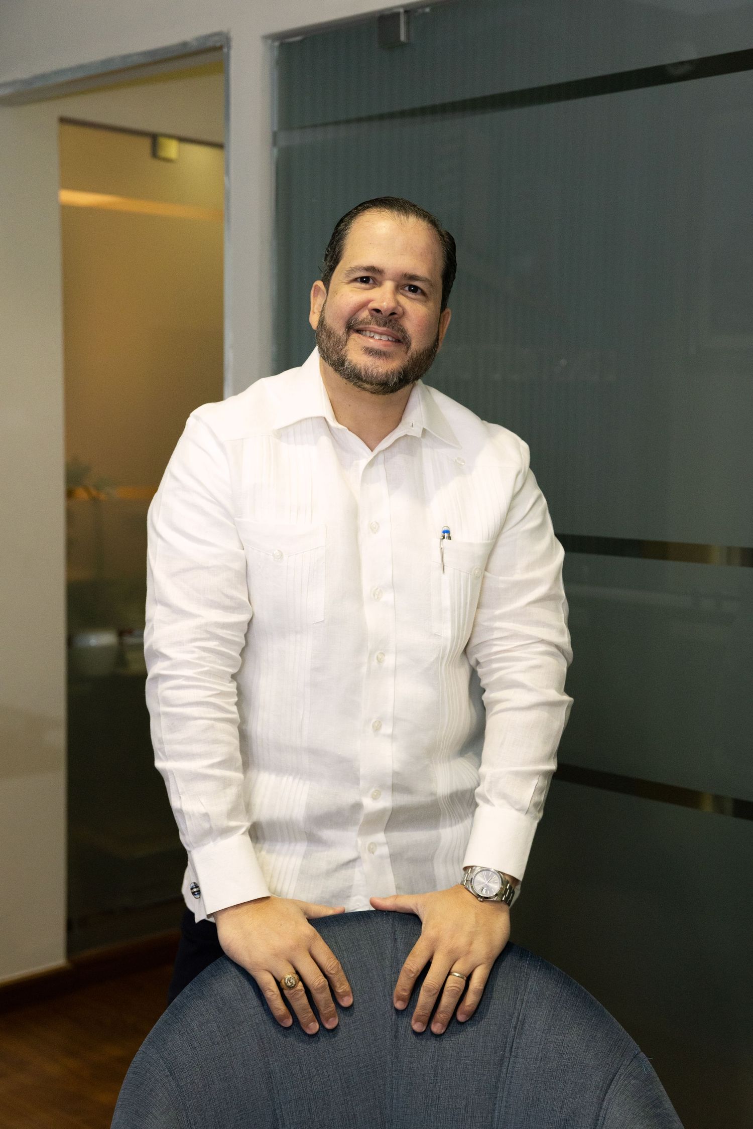 Smiling man in white guayabera shirt seated casually in modern office, wearing a watch and jeans.