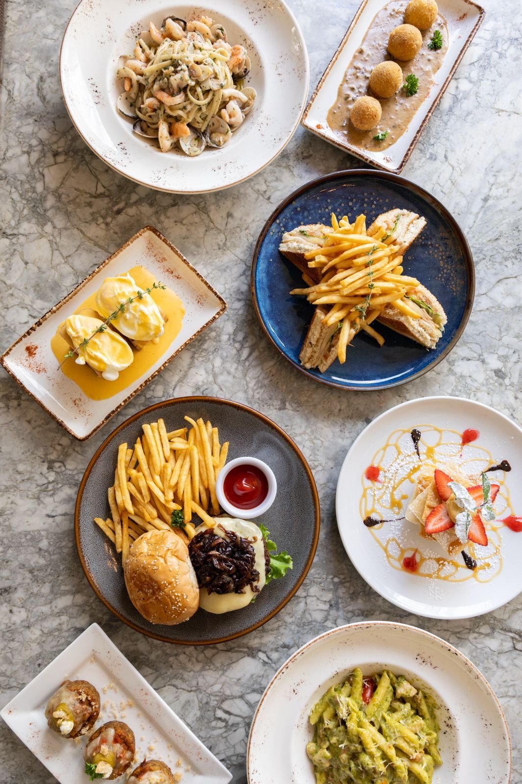 Overhead view of a restaurant spread featuring pasta, burger with fries, sandwich, dessert, and appetizers on marble table.