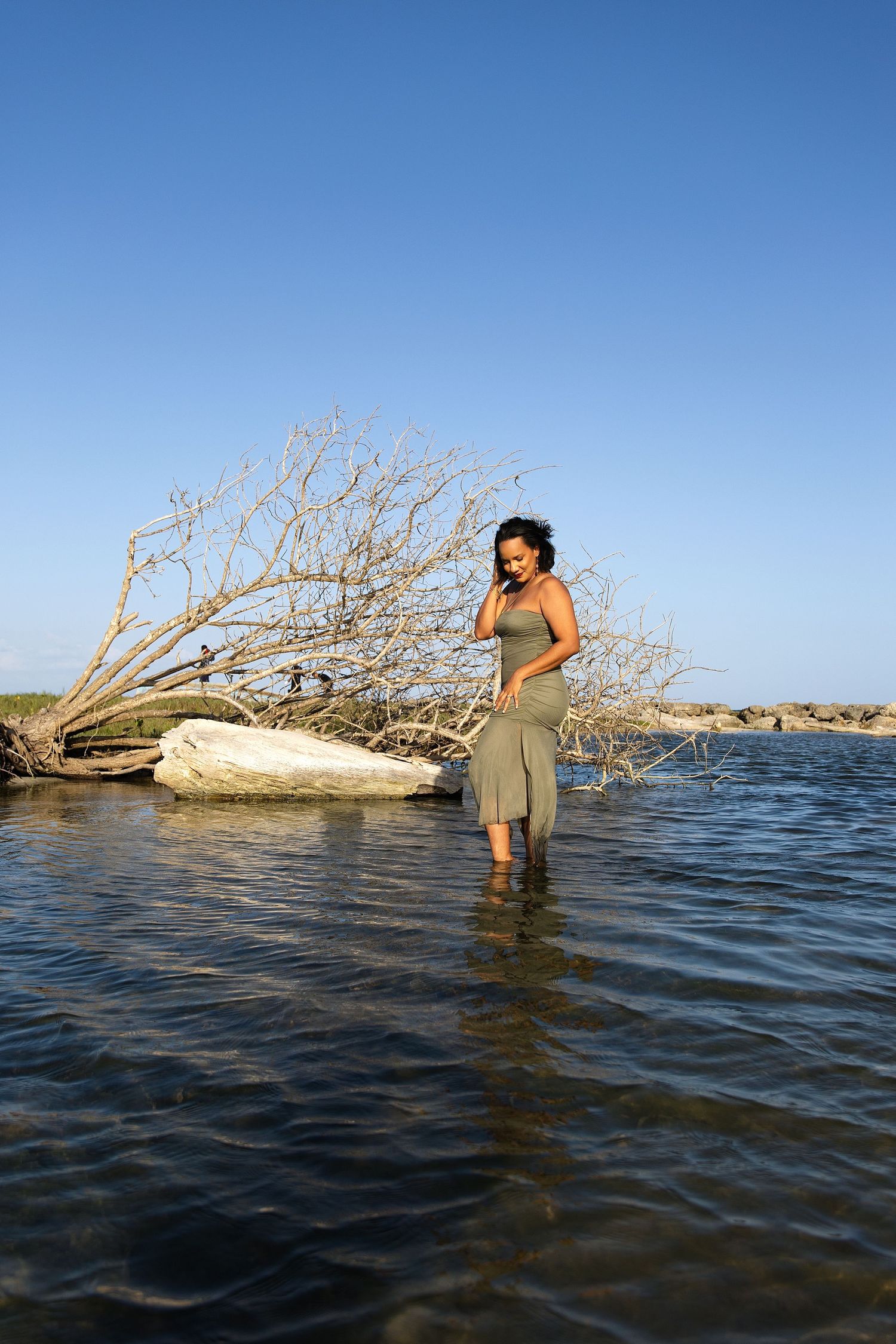 A woman in a gray dress stands wading in shallow water near driftwood under a clear blue sky.