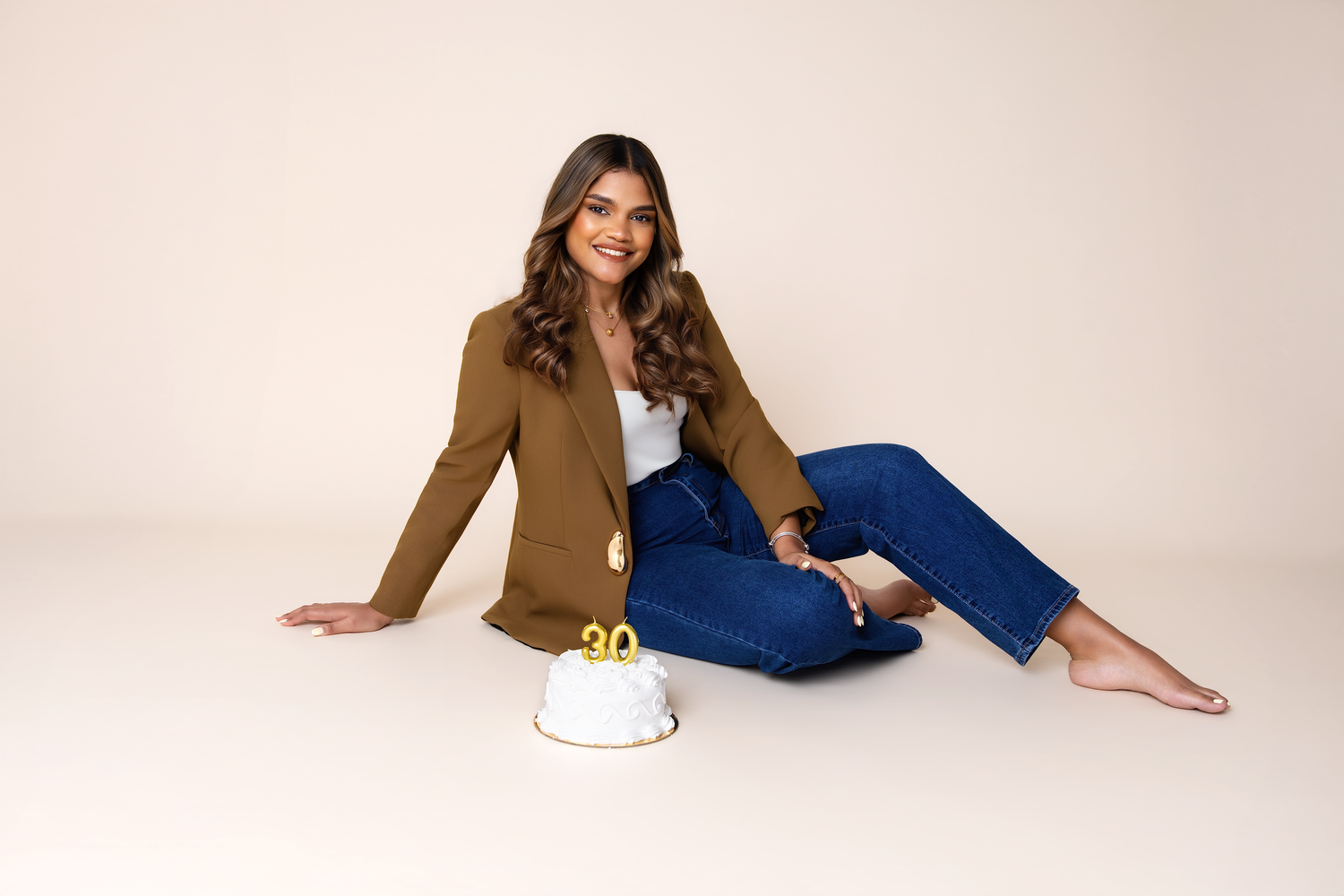 Woman in brown blazer and jeans sits beside a white birthday cake with gold number candles on a neutral background.