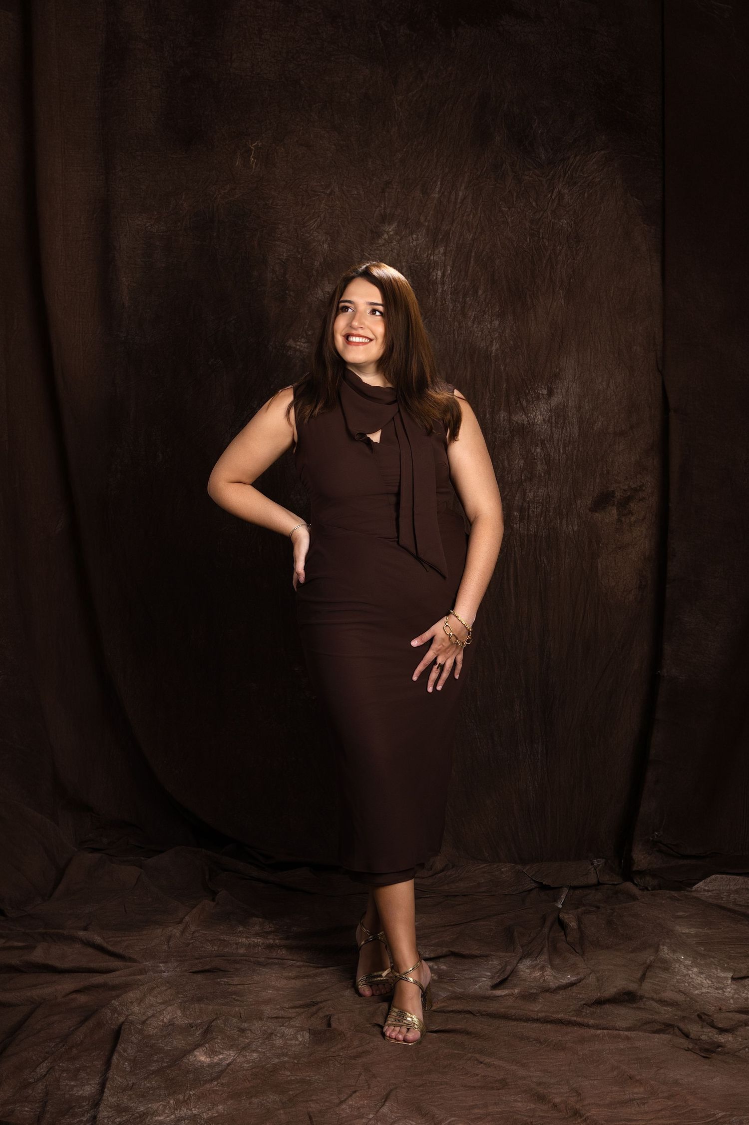 Woman in brown sleeveless midi dress posing confidently against a dark brown textured studio backdrop.