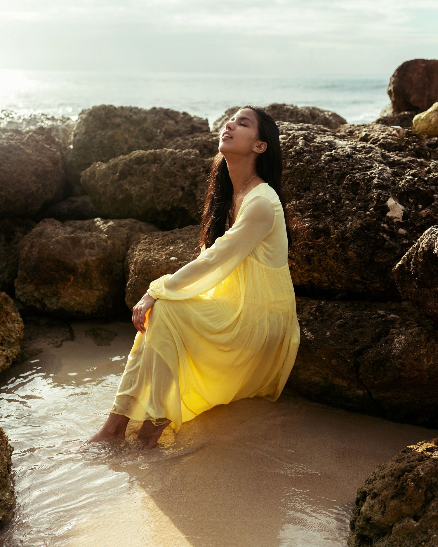 A serene beach portrait featuring a flowing yellow dress against coastal rocks at sunset.
