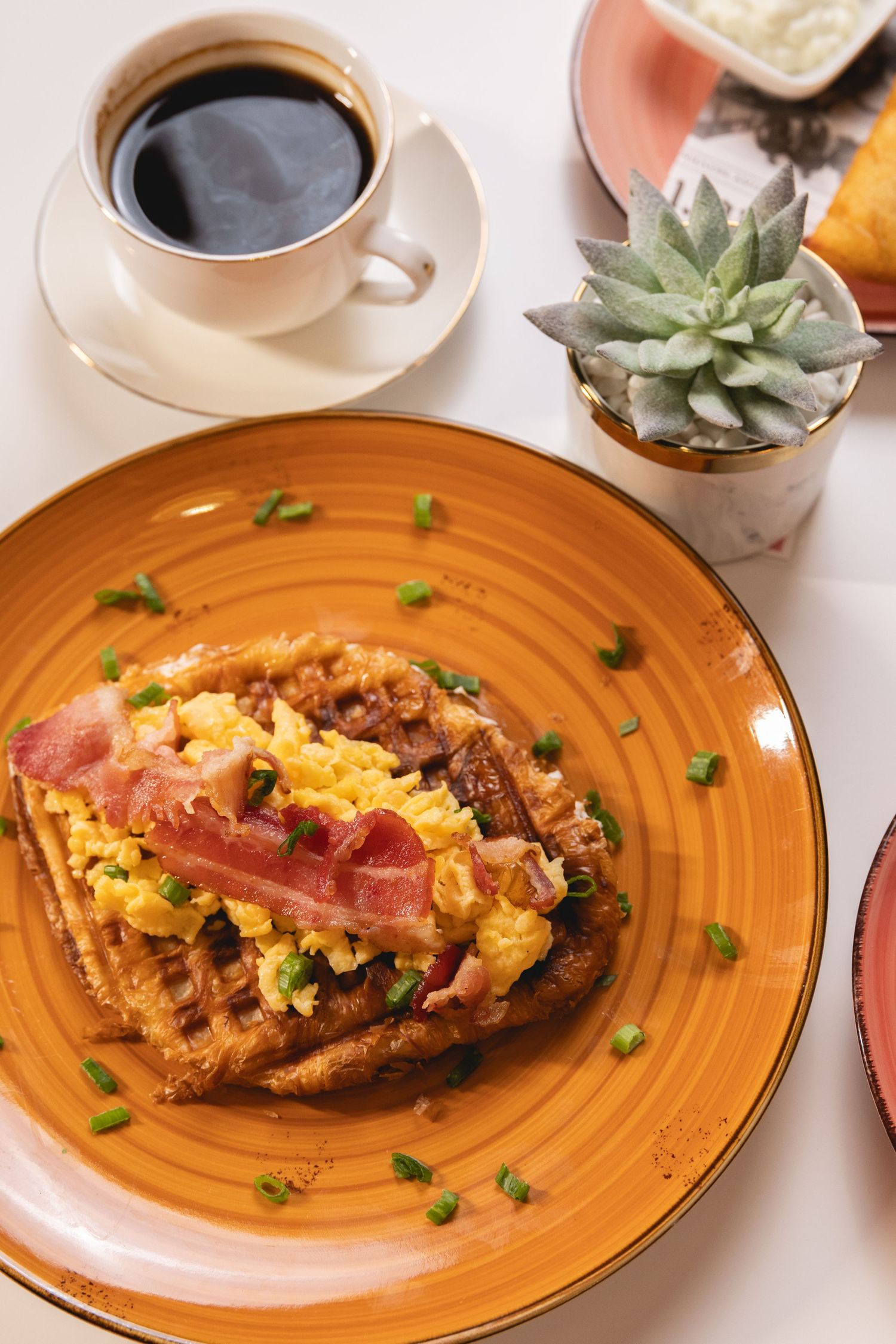 Savory breakfast waffle topped with scrambled eggs and crispy bacon on an orange plate, served with black coffee.