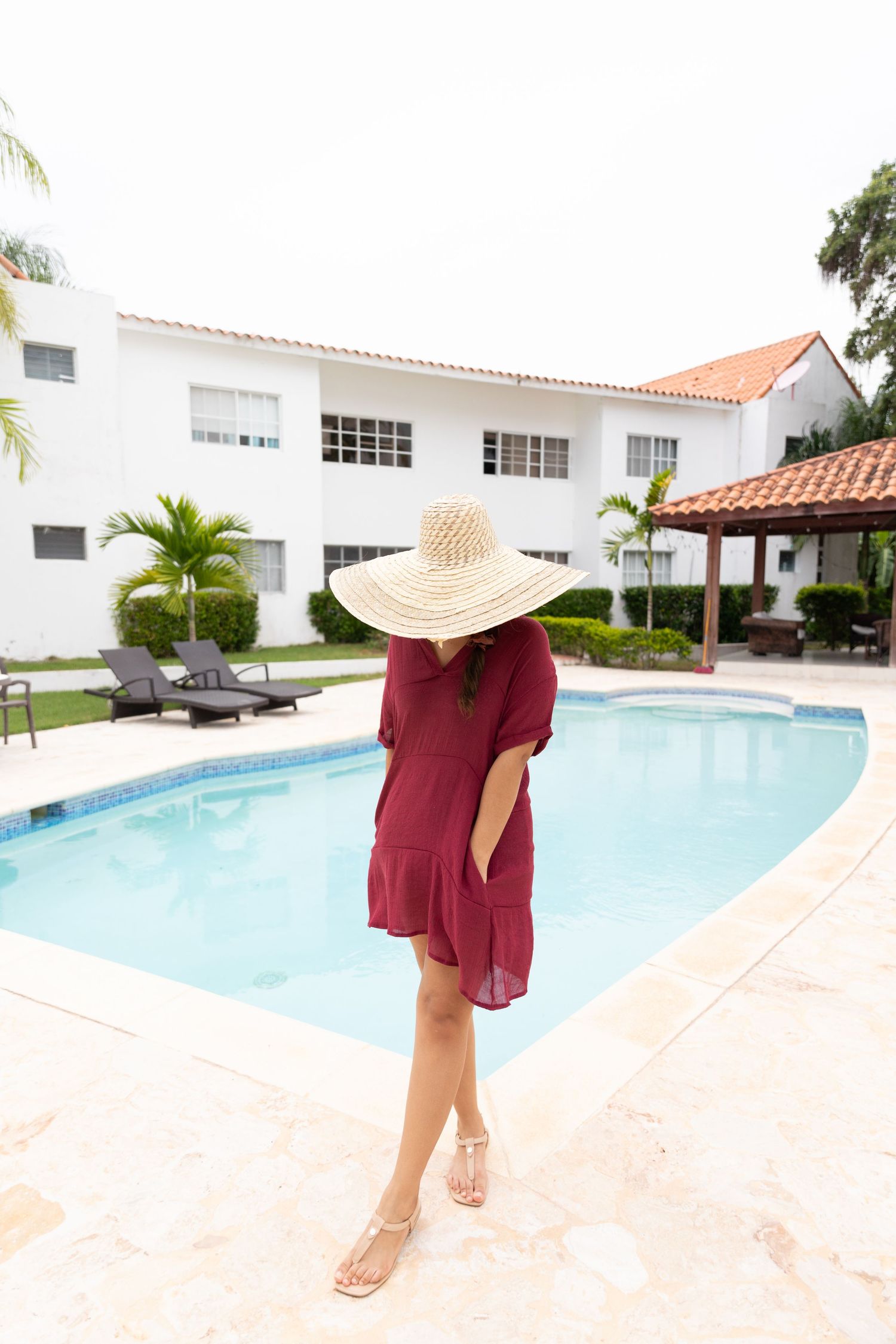 Woman in burgundy dress and wide-brim straw hat standing by a resort pool with white building backdrop.
