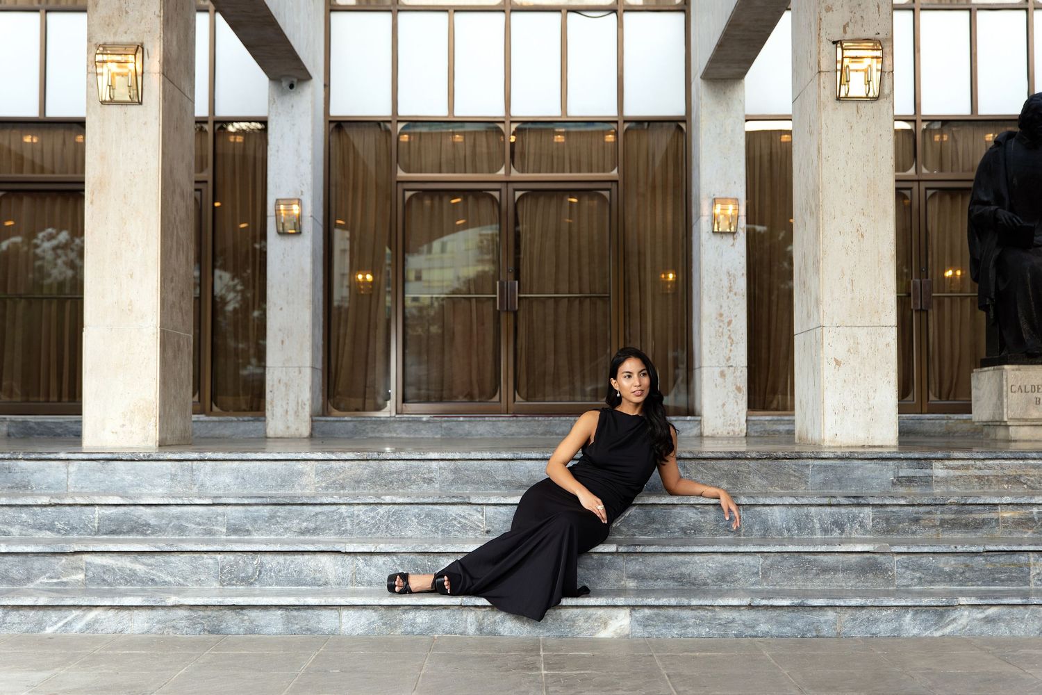 Woman in black outfit posing on marble steps outside elegant building with ornate wooden doors.