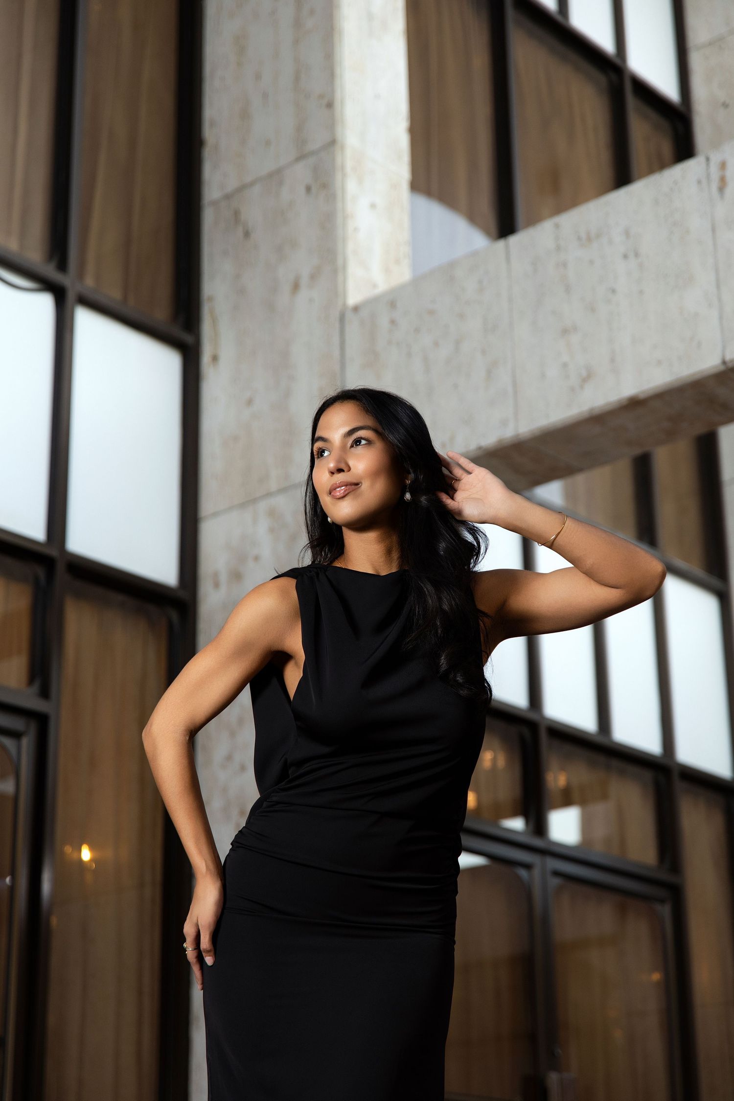 Woman in black sleeveless dress poses confidently in front of modern building with large windows.