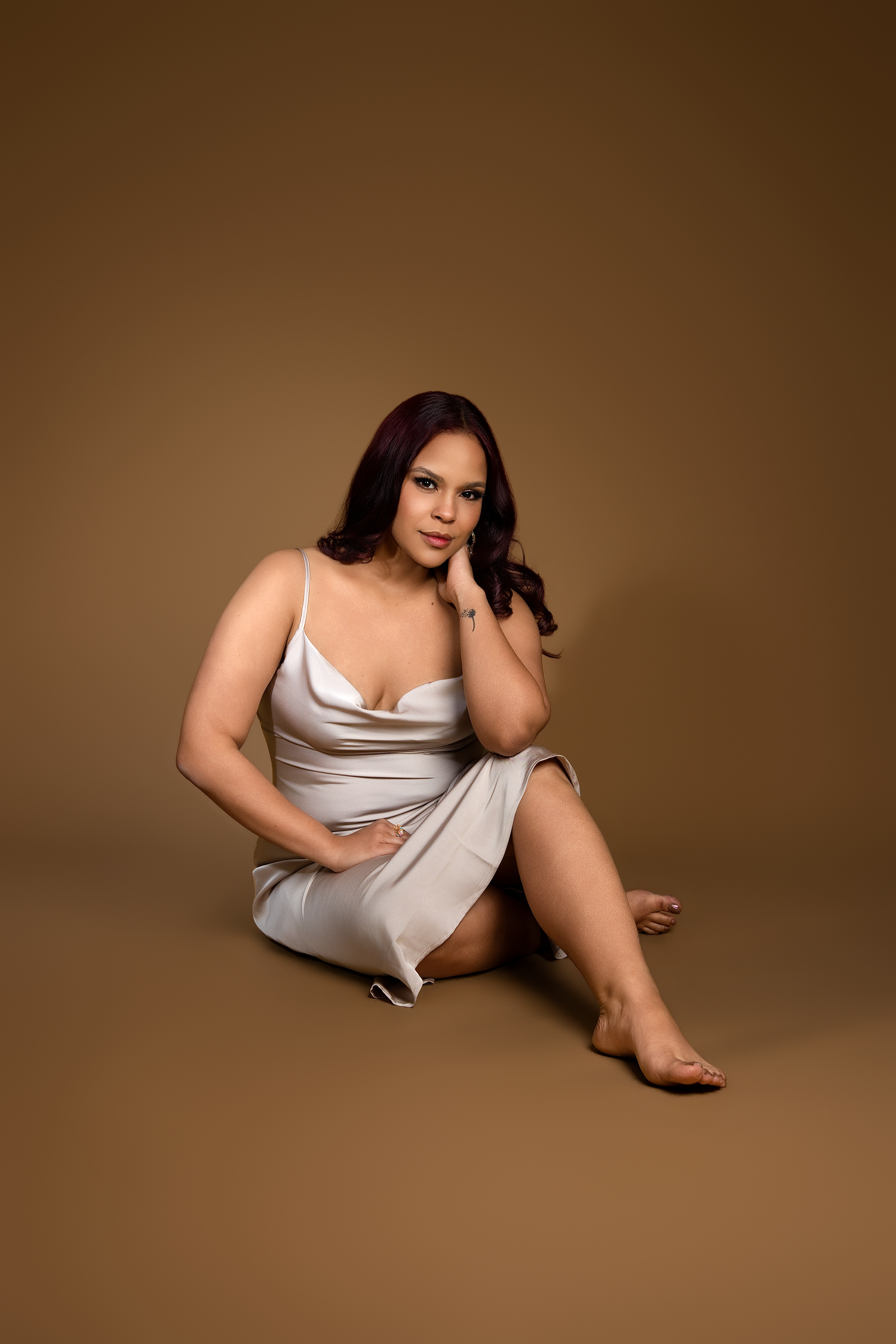 A plus-size woman in a silver slip dress sits gracefully on a brown studio backdrop, resting her chin on her hand.