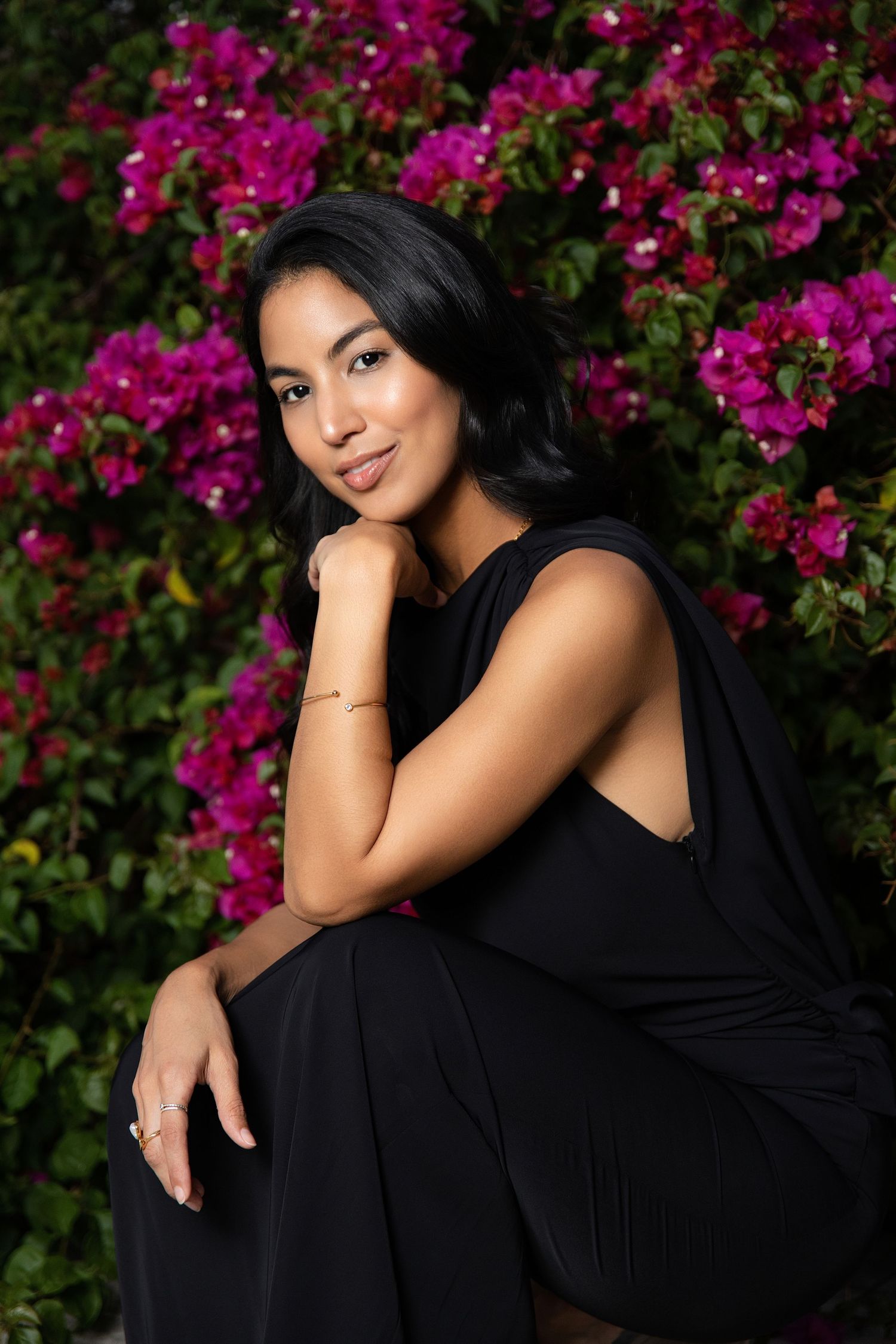 A young woman in a black dress sits gracefully in front of vibrant pink bougainvillea flowers, smiling softly.