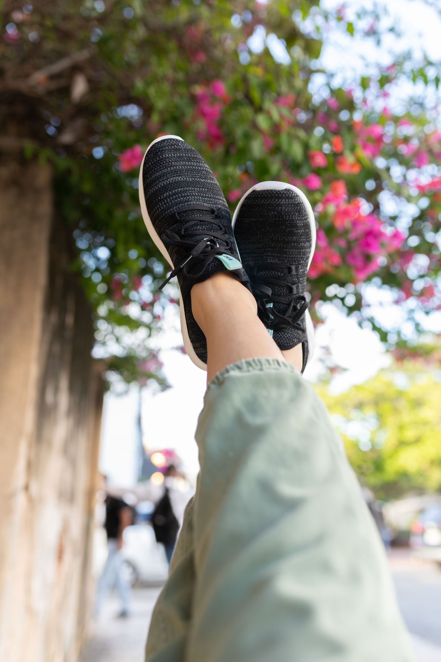 Person wearing black knit sneakers and olive green pants with legs raised against a backdrop of pink flowers and trees.
