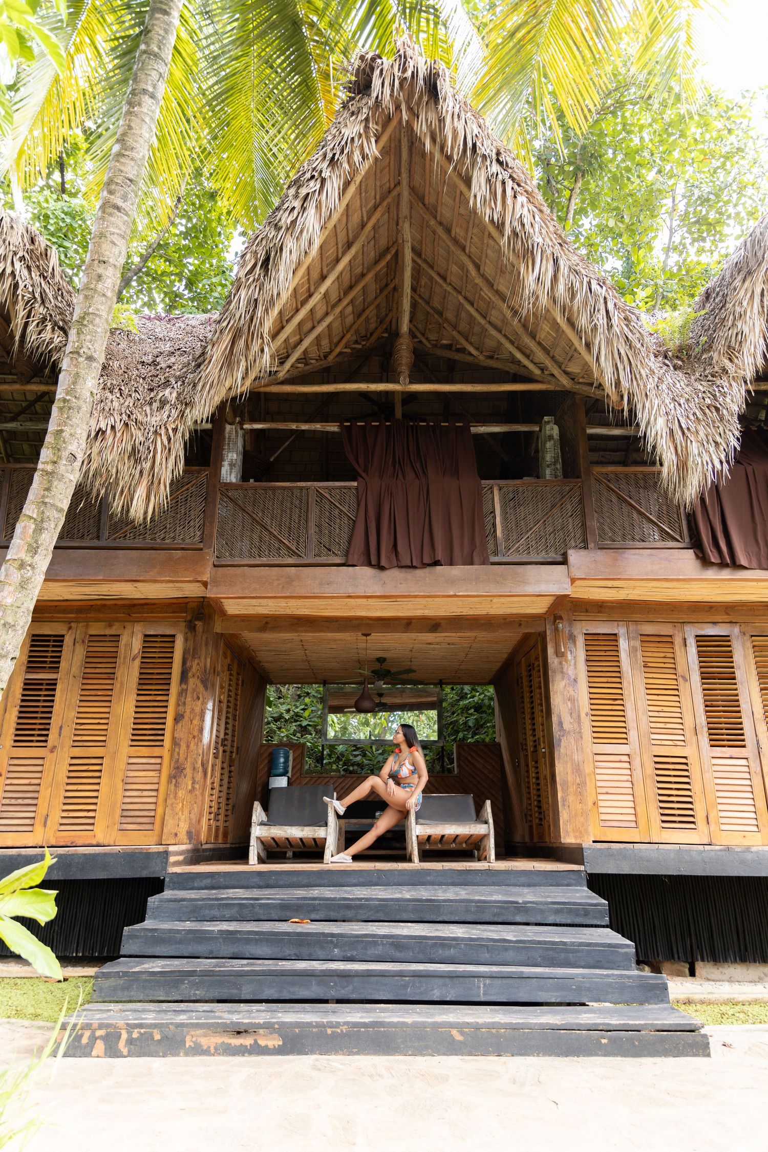Woman relaxing on lounge chair at tropical wooden bungalow with thatched roof surrounded by palm trees.