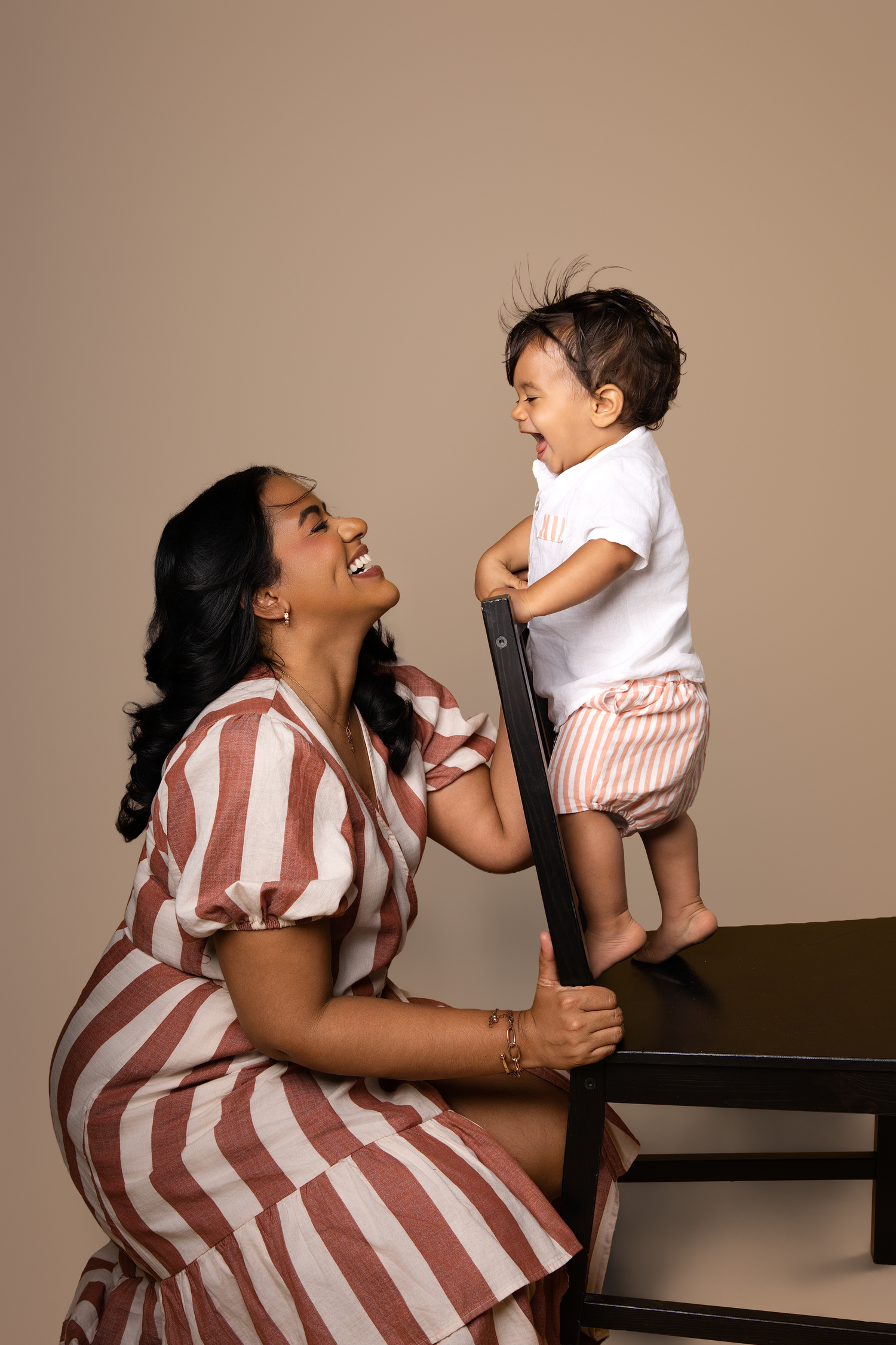 A mother in a striped dress laughs joyfully with her happy baby standing on a dark table against a beige background.