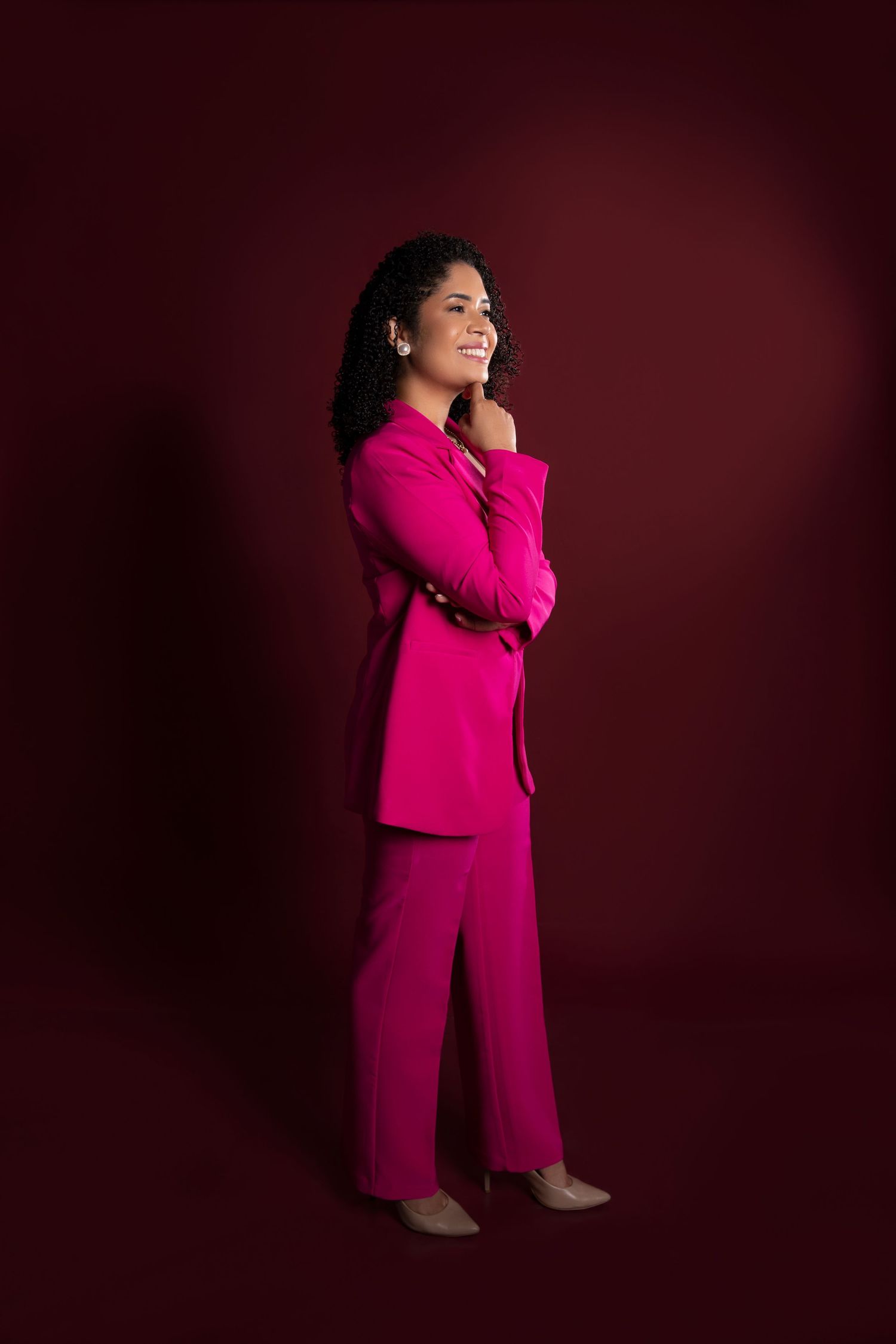 Person in vibrant fuchsia pantsuit poses confidently against dark burgundy backdrop.