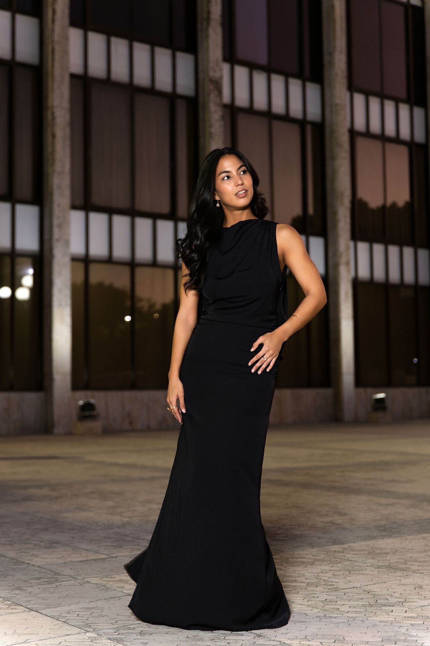 Woman in elegant black sleeveless floor-length gown posing outdoors at night in front of a modern building.