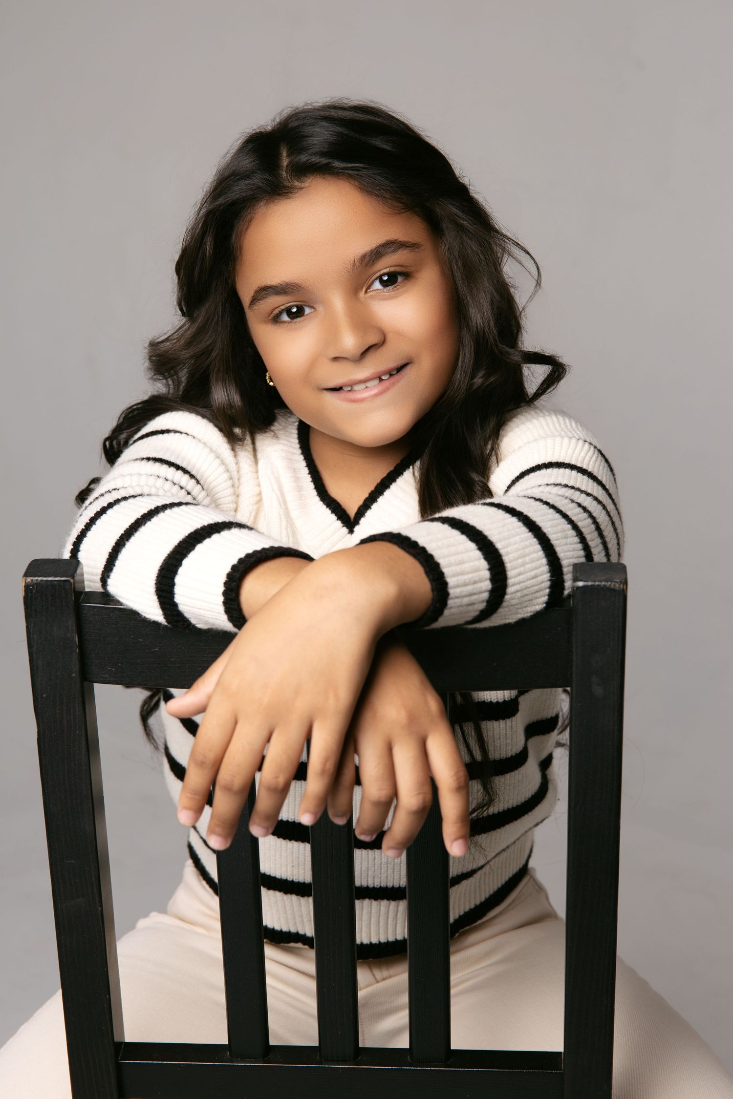 Child in striped sweater posing playfully with black chair against grey background.