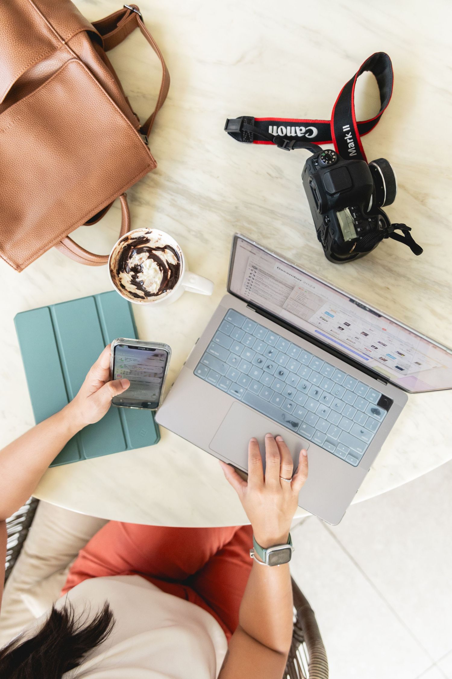 Overhead view of woman using laptop and smartphone at cafe table with Canon camera, coffee, tablet, and brown leather bag.