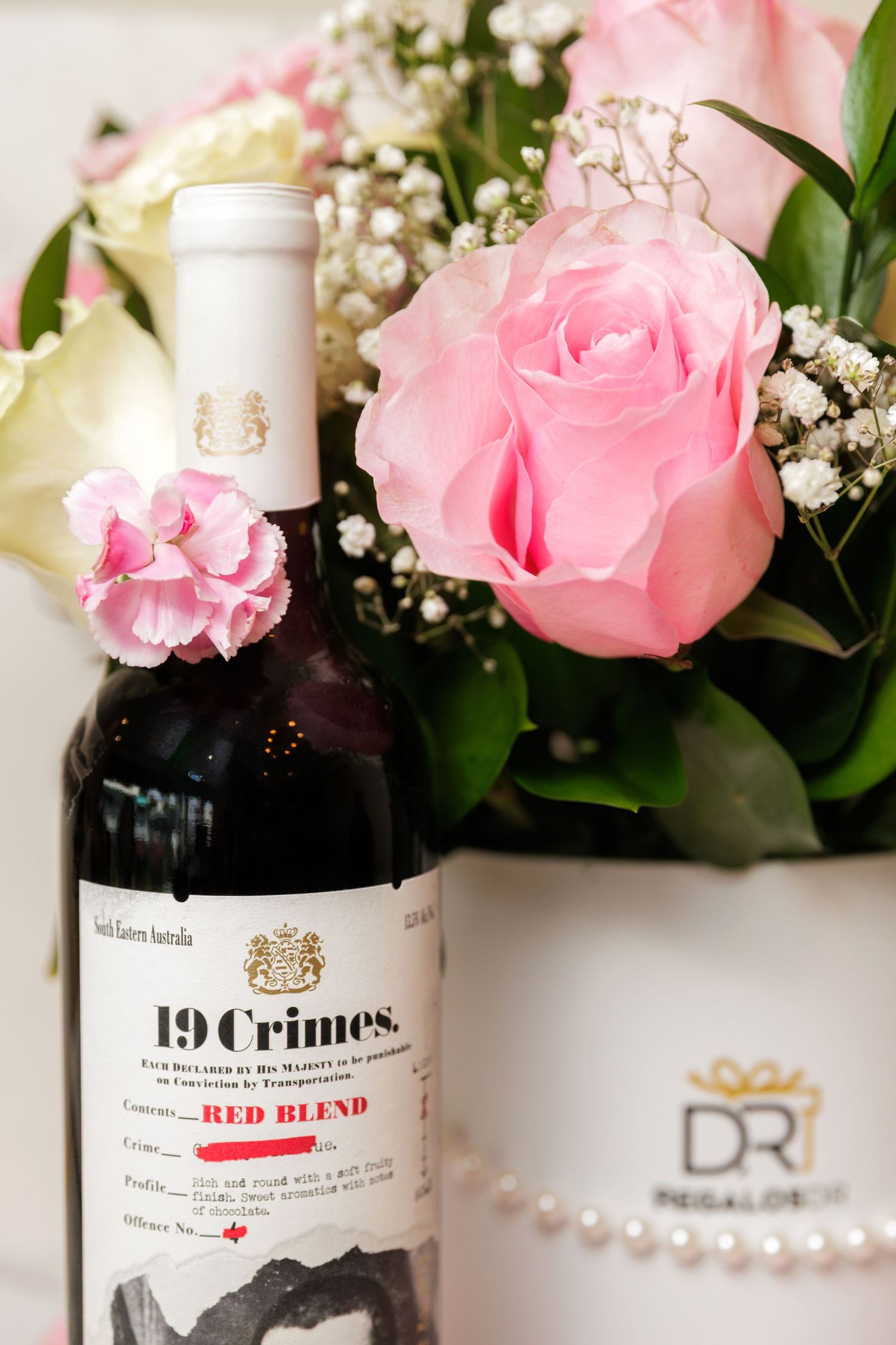 19 Crimes Red Blend wine bottle surrounded by pink roses and white flowers in a DR Regalos gift box.