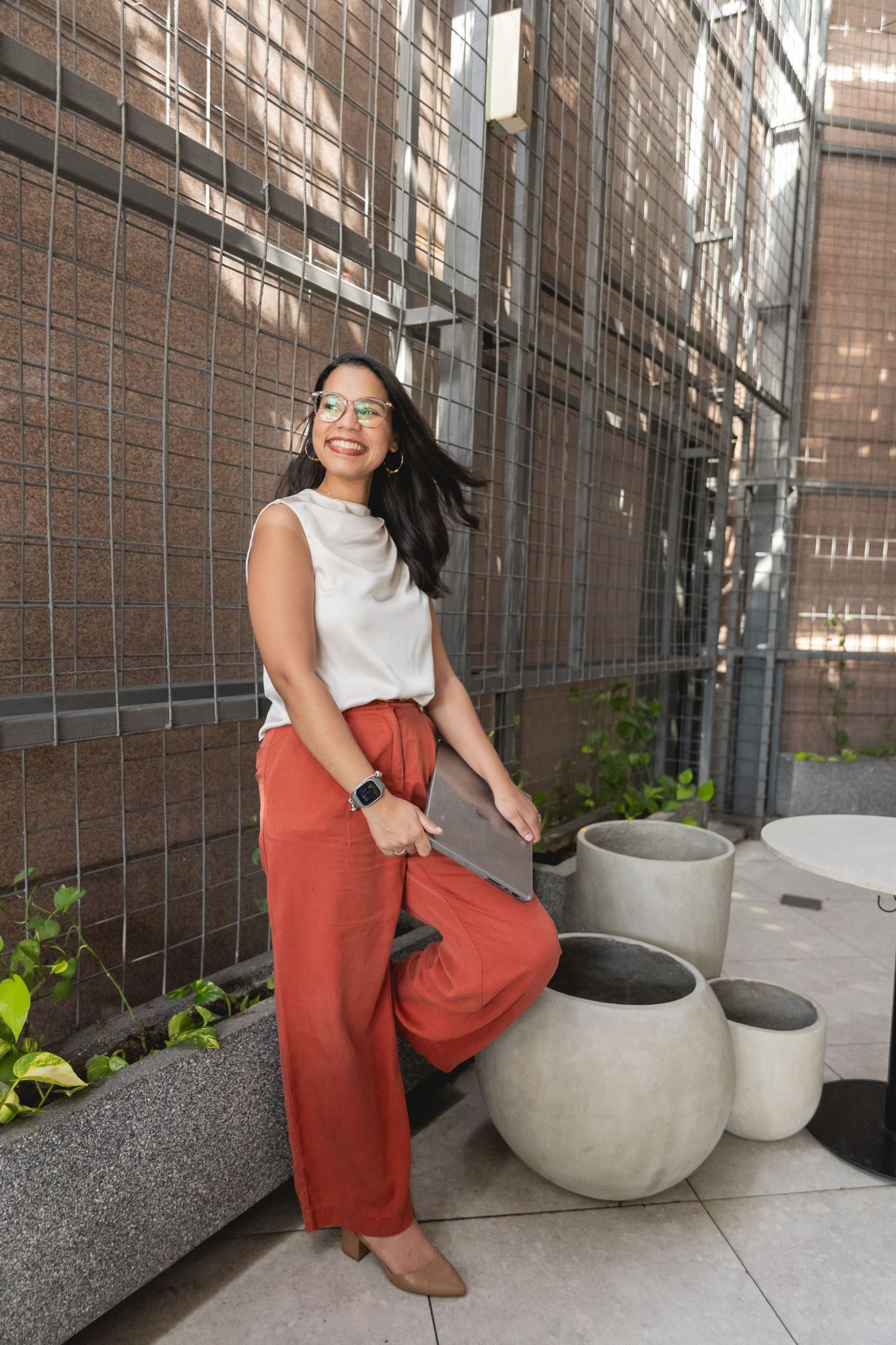 Woman in white top and rust wide-leg pants sits outdoors holding a laptop, smiling near concrete planters.