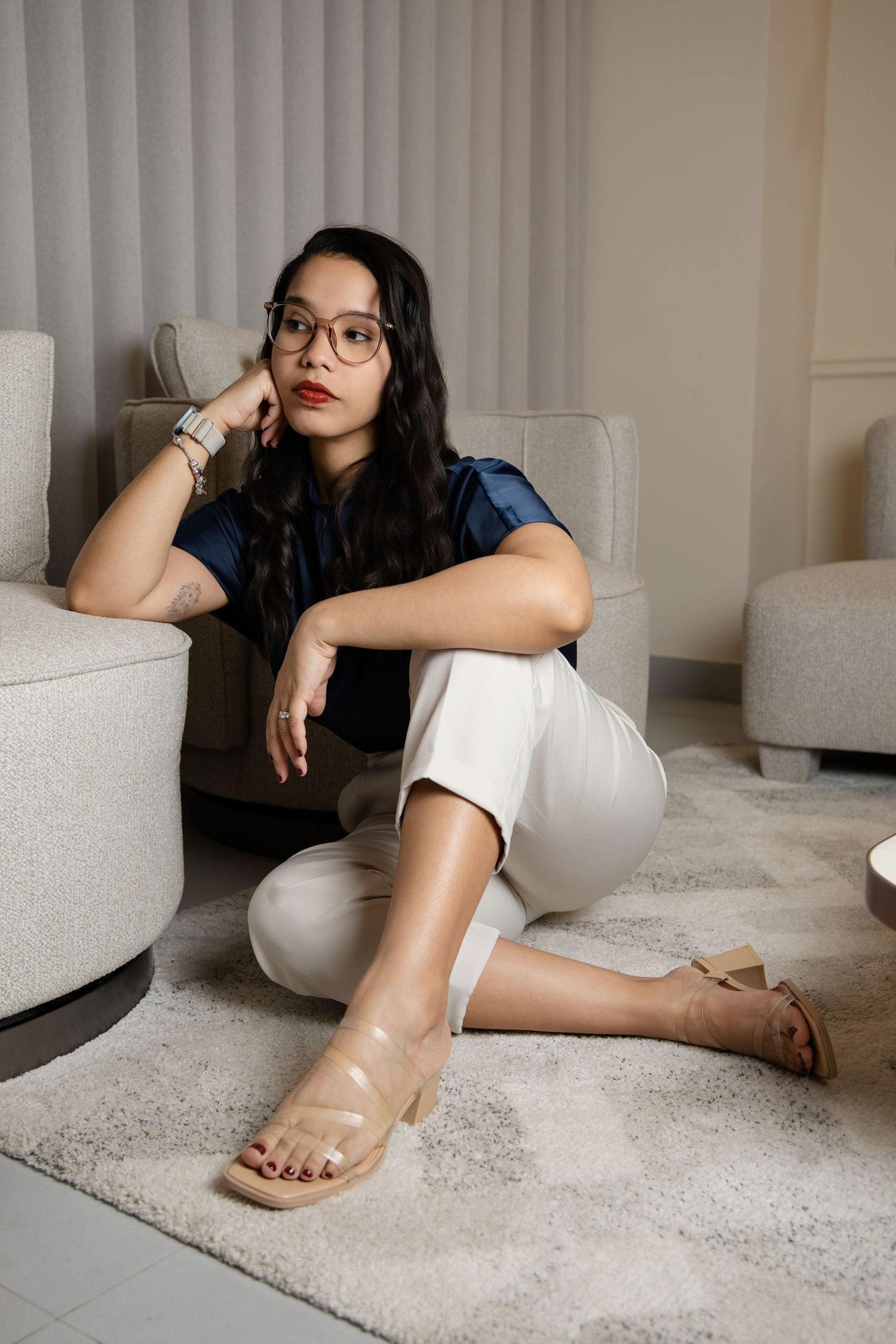Woman in glasses, blue top, white pants, and sandals sitting on a carpet leaning against a chair indoors.