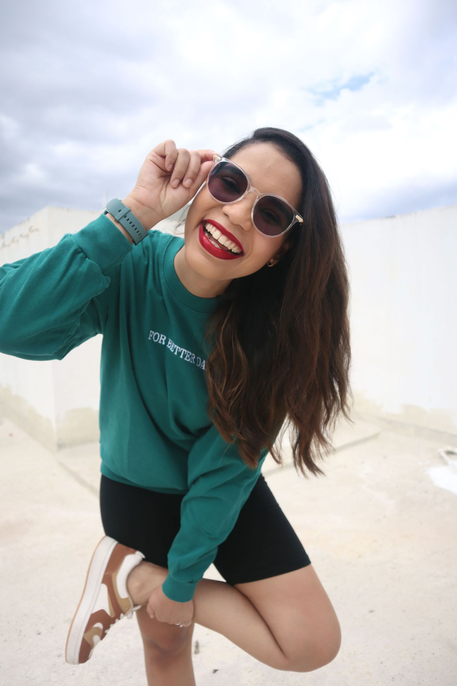 Smiling woman in green sweatshirt, black shorts, and sunglasses posing outdoors with red lipstick and long dark hair.