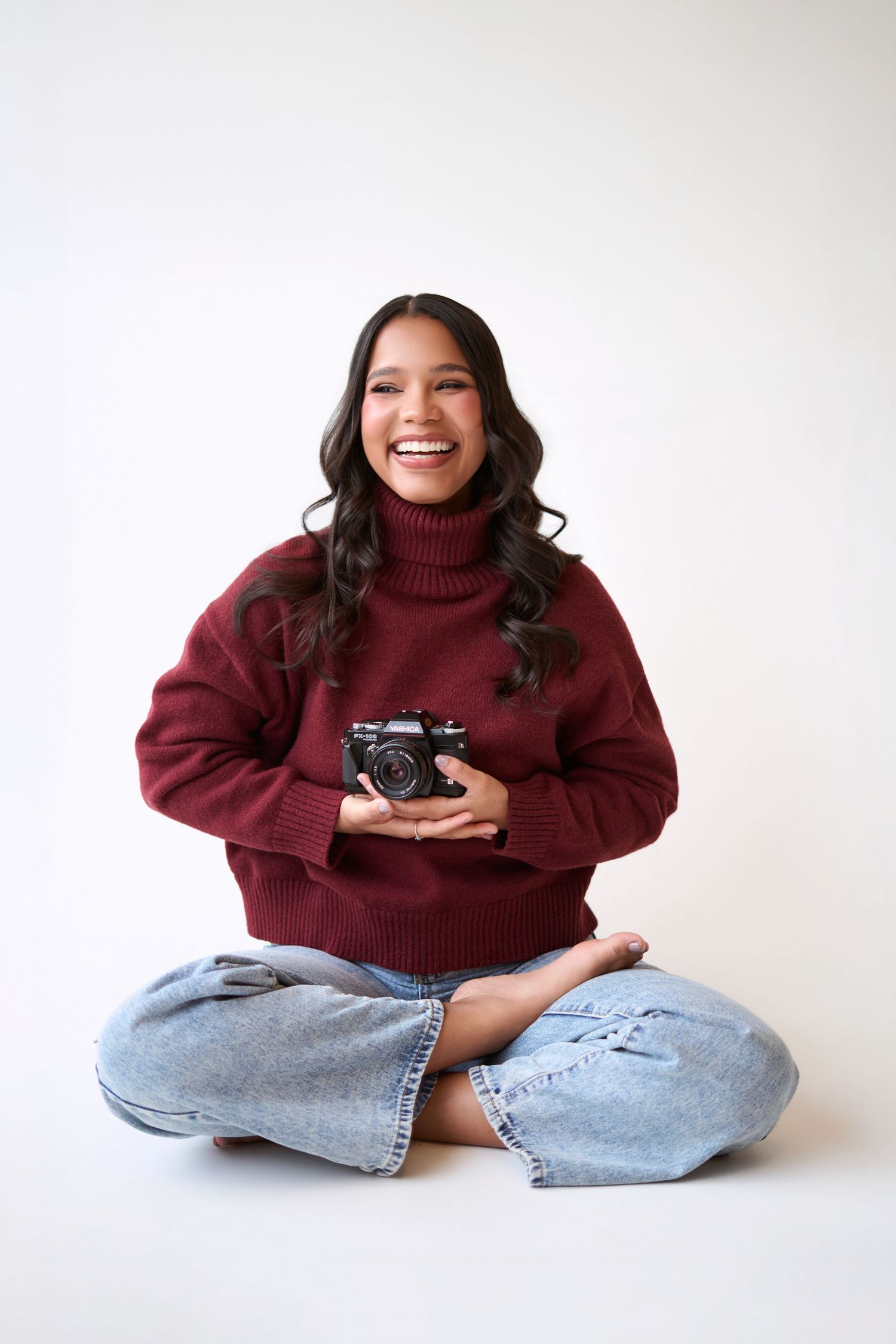 Smiling young woman sitting cross-legged holding a film camera, wearing a burgundy turtleneck sweater and jeans.
