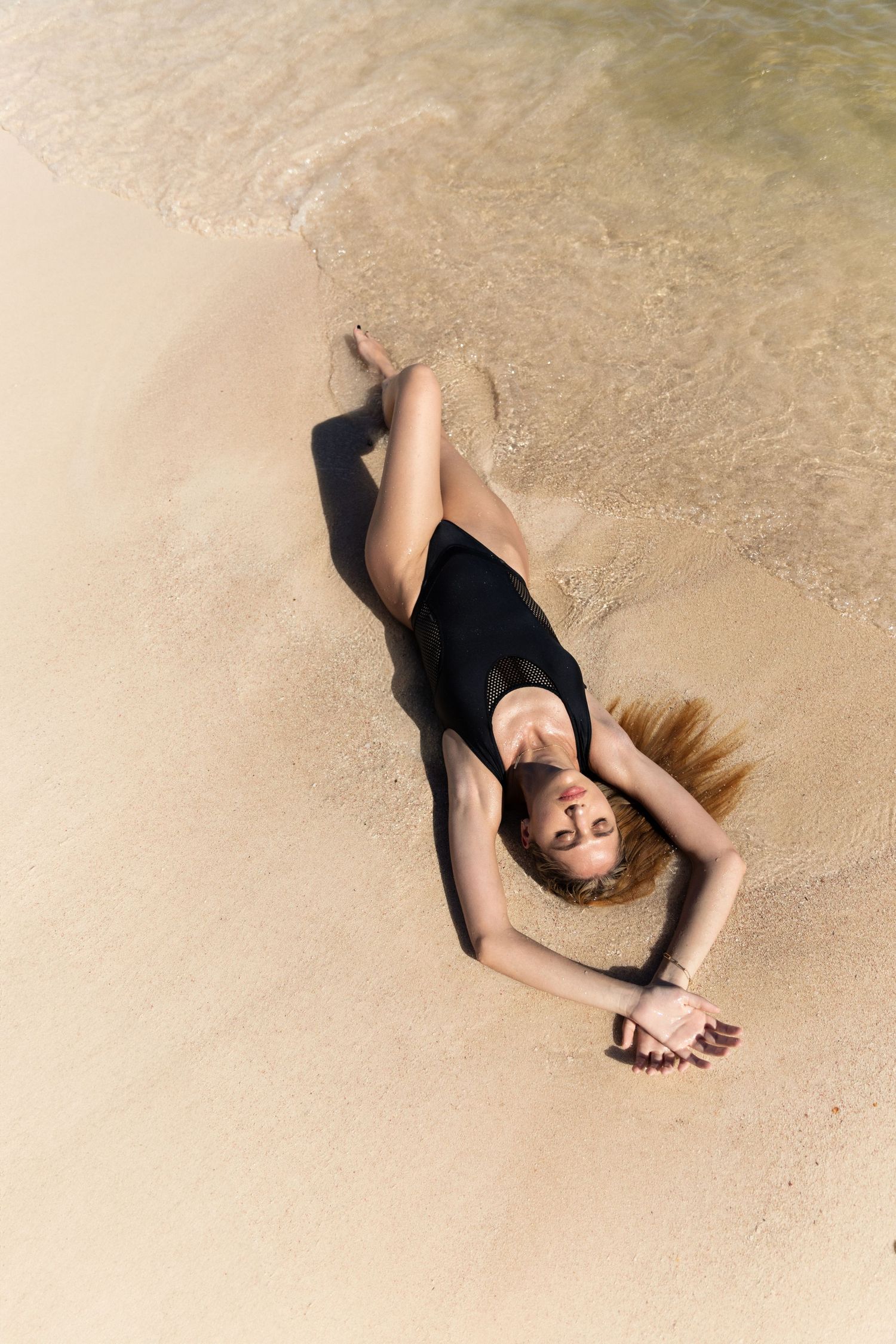 Person in black swimsuit lying on sandy beach as waves wash over.