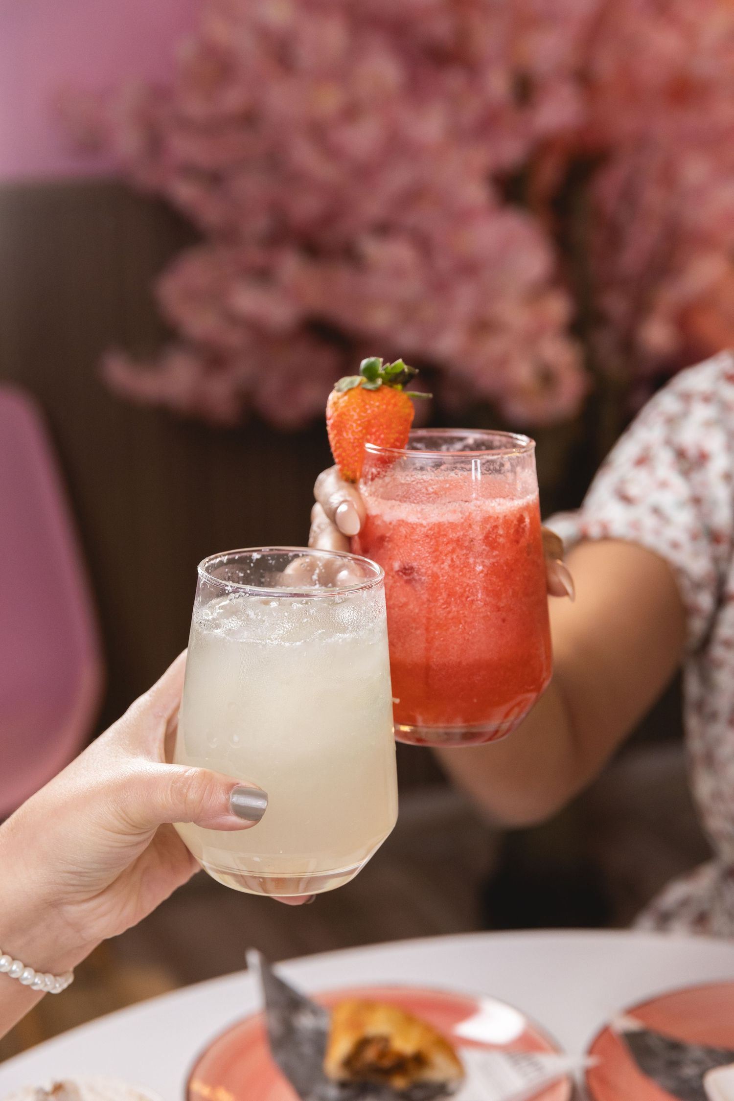 Two hands clinking glasses with a white lemonade and a strawberry garnished pink cocktail at a restaurant.