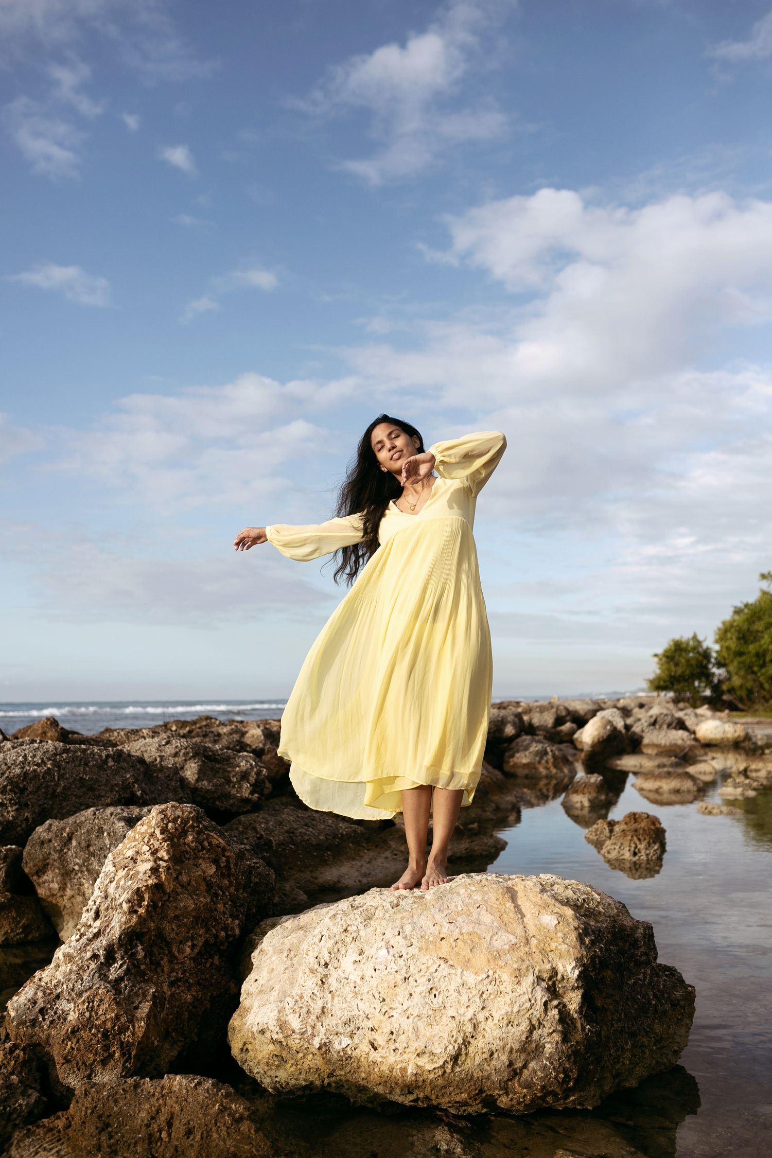 Woman in yellow dress poses gracefully on large coastal rocks during sunset at the beach.
