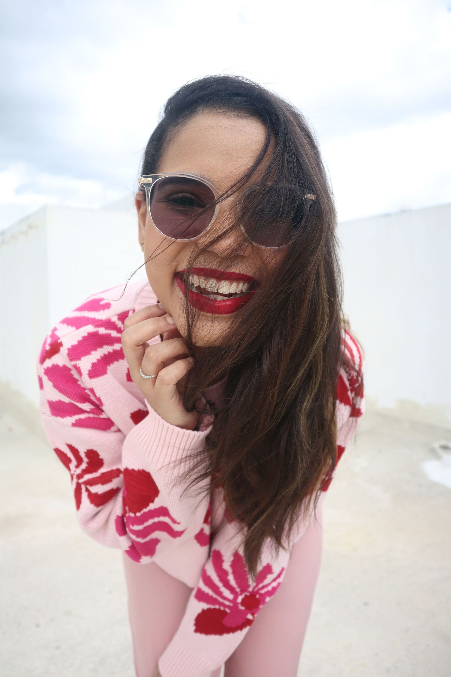 Smiling woman in pink floral sweater and round sunglasses laughing outdoors with red lipstick and windswept hair.