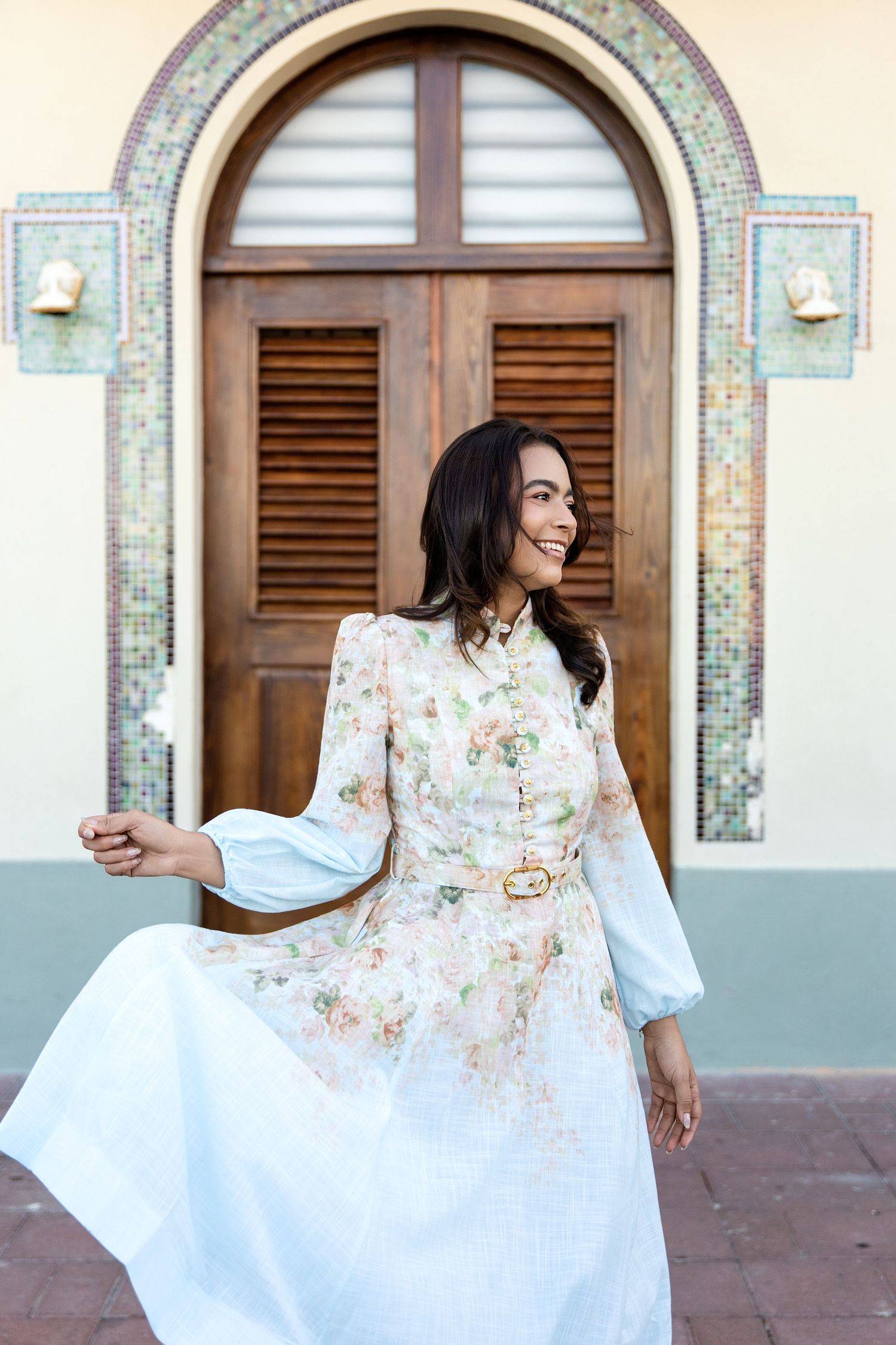 Woman in floral white belted midi dress twirling outdoors near ornate arched wooden door with mosaic tile accents.