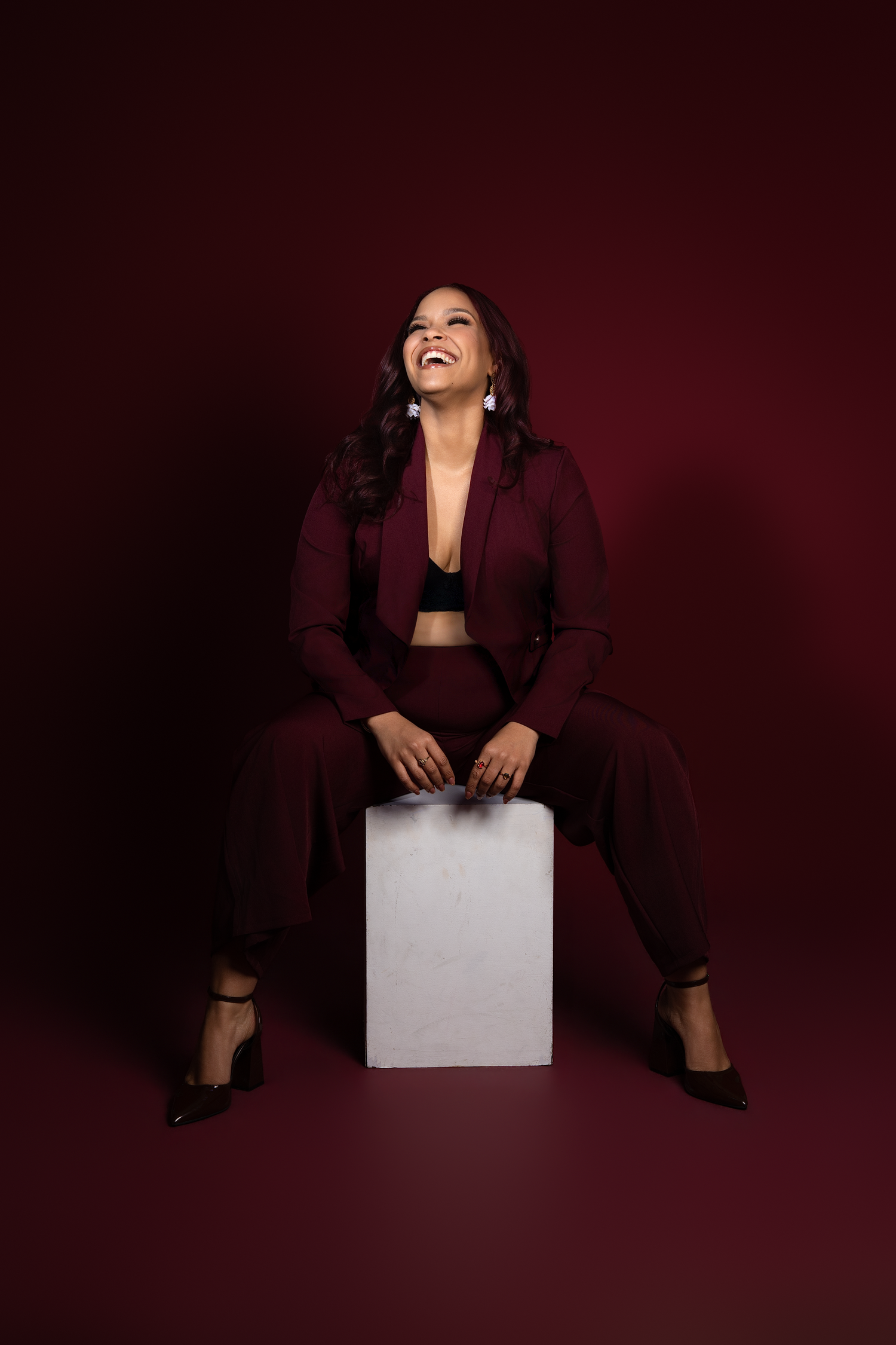 A woman in a burgundy suit sits on a white cube, laughing joyfully against a dark red background.