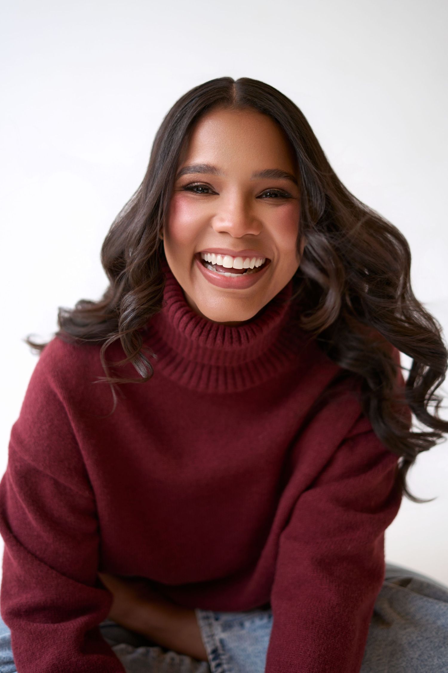 A young woman with wavy dark hair smiles brightly, wearing a burgundy turtleneck sweater against a white background.