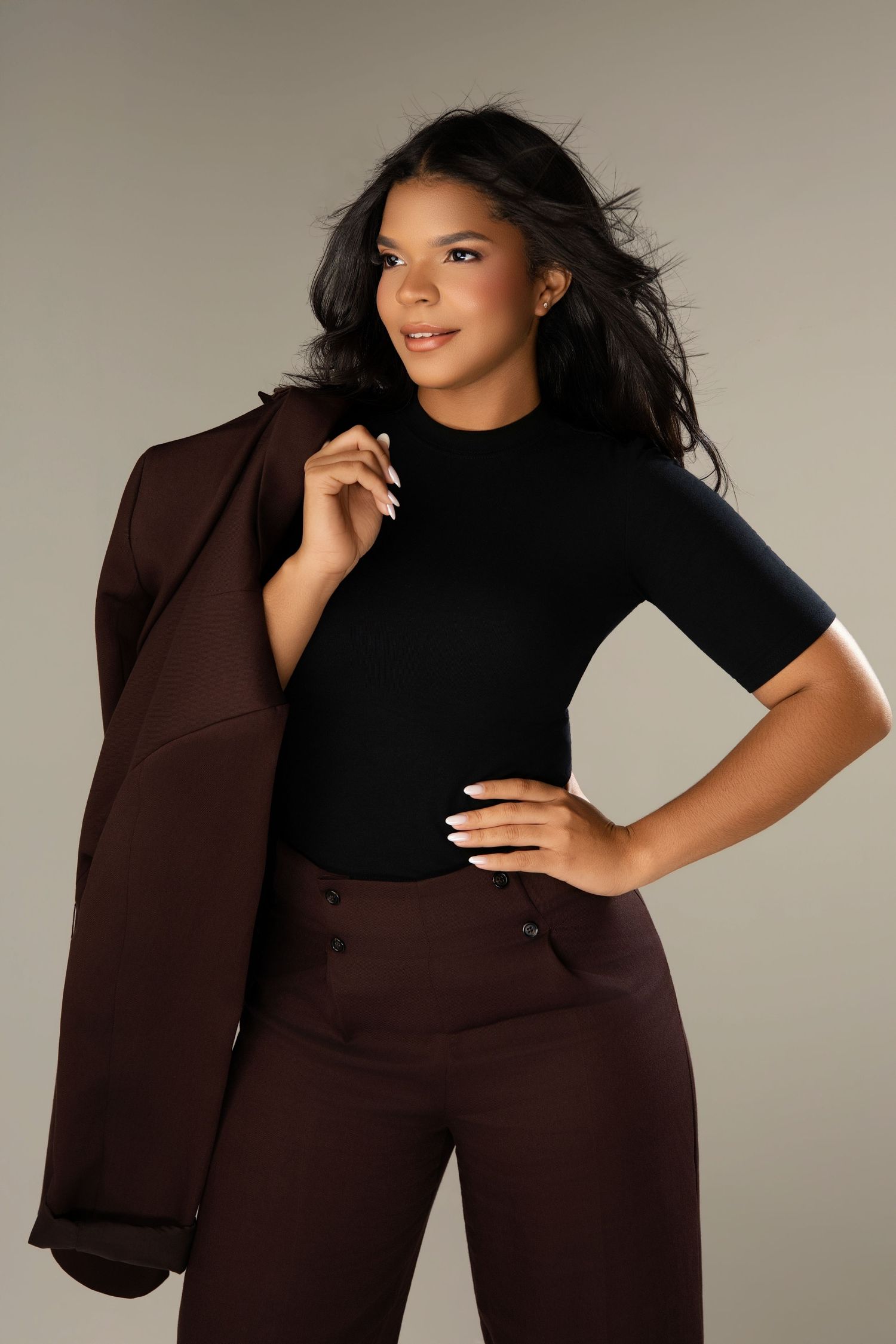 Woman in black turtleneck top and brown trousers holding a blazer over her shoulder in a studio setting.