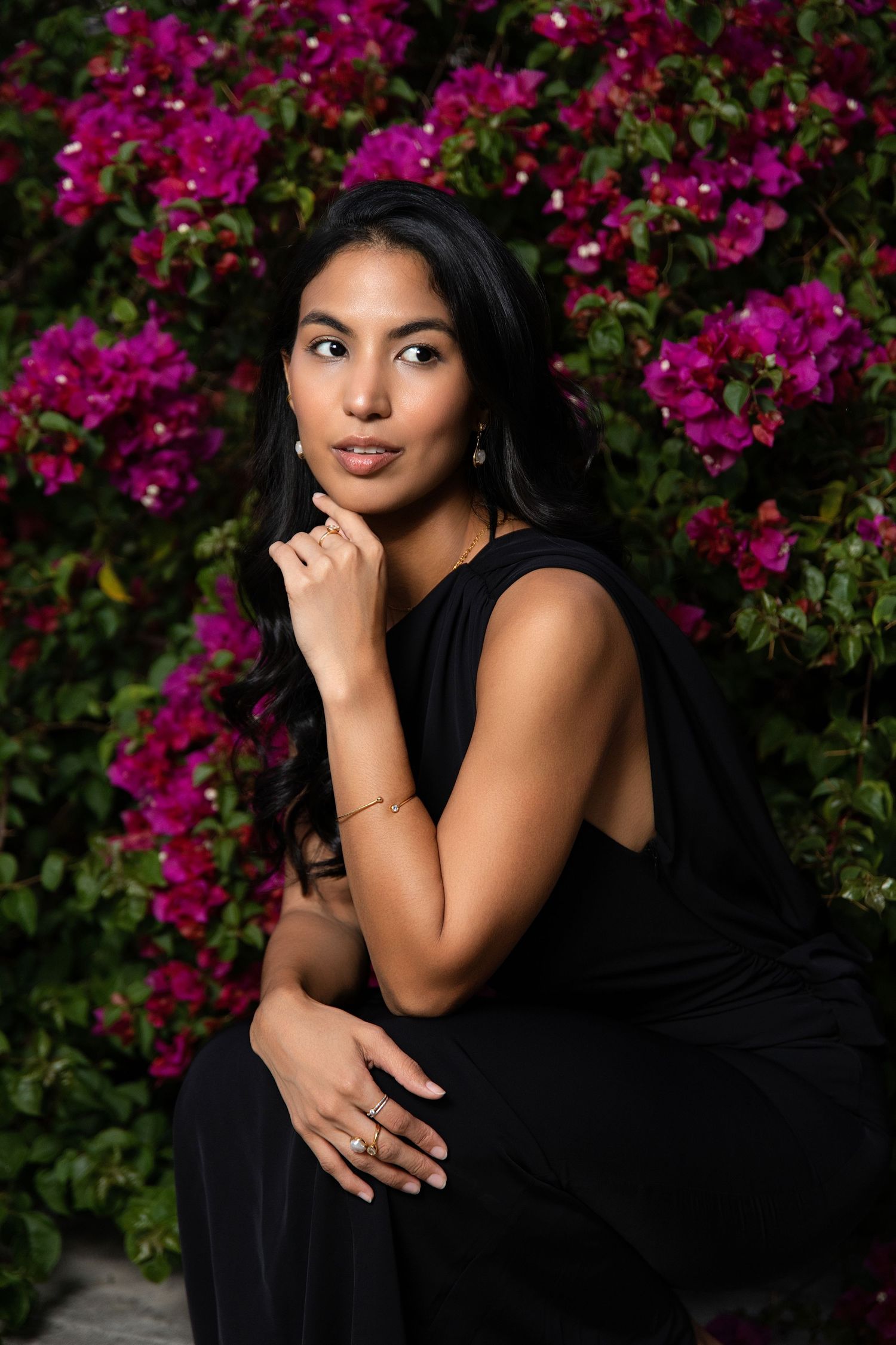 Woman in black dress poses thoughtfully before vibrant pink bougainvillea flowers, wearing gold jewelry.