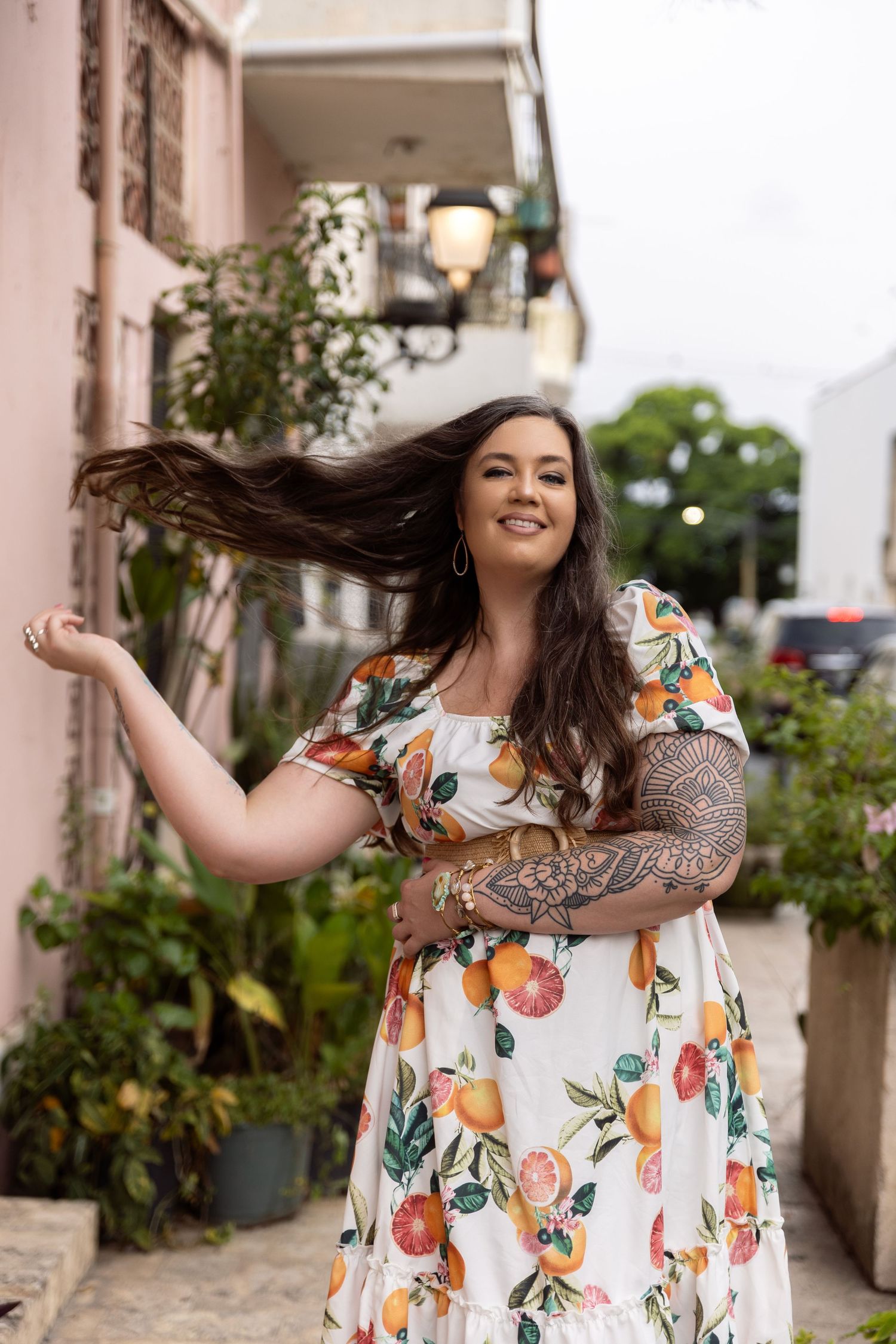Woman in white floral dress with tattoo sleeve twirls playfully on urban street.