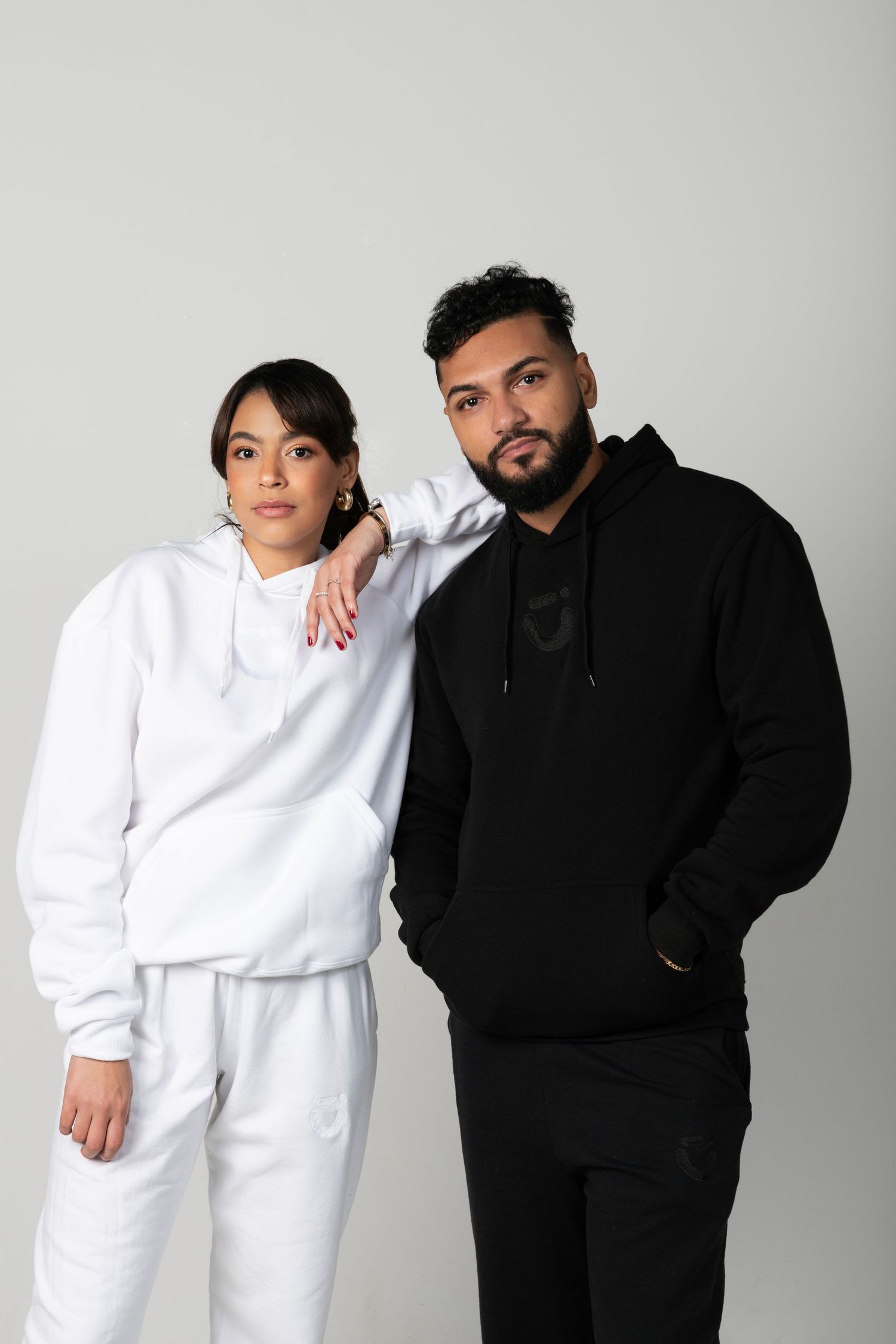 Two people in contrasting black and white hoodies pose together against grey background.