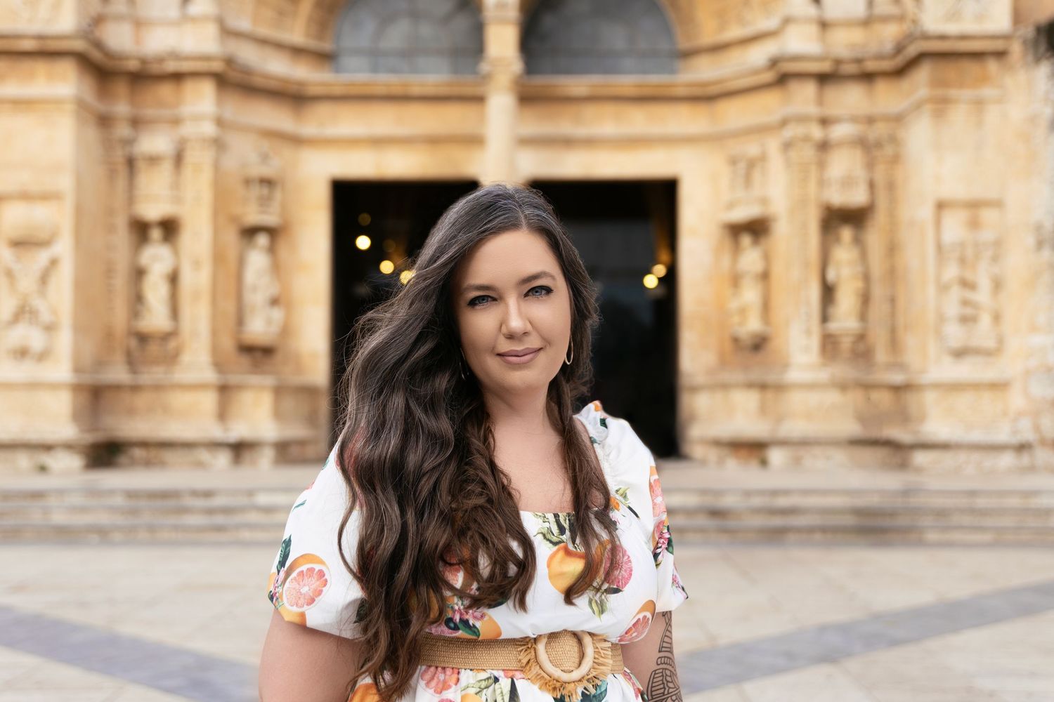 Woman with long dark hair wearing floral dress poses in front of ornate historic building.