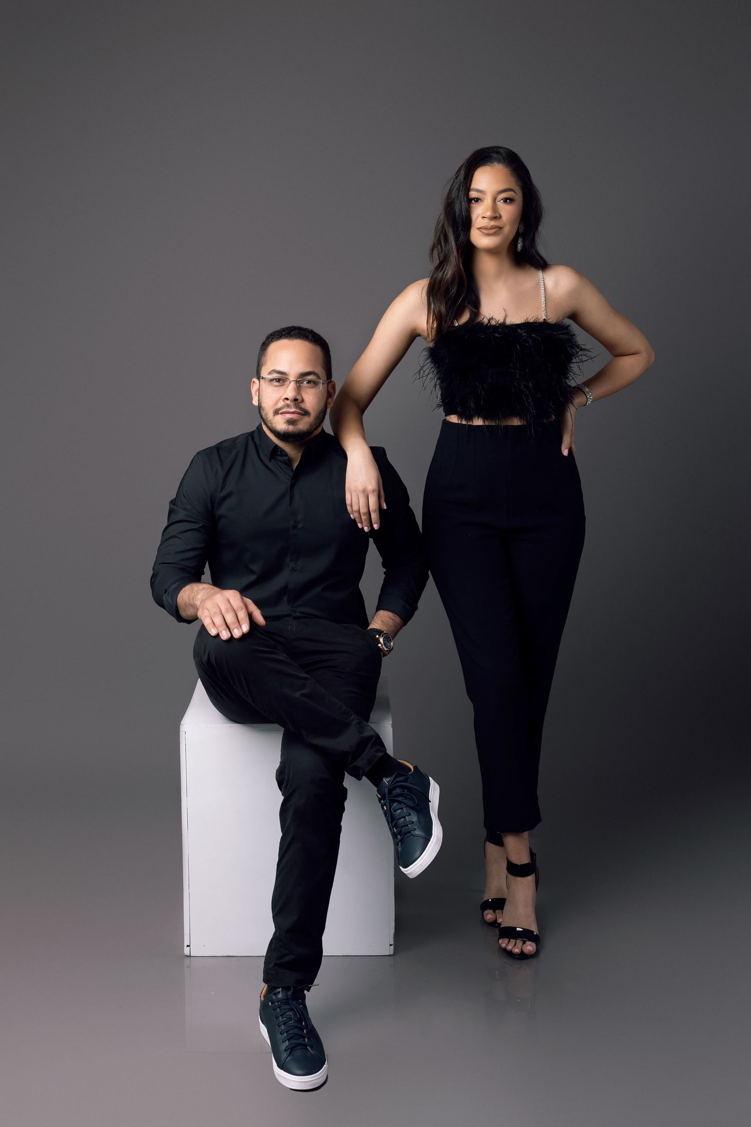 Couple in matching black outfits posing for studio portrait against gray background.