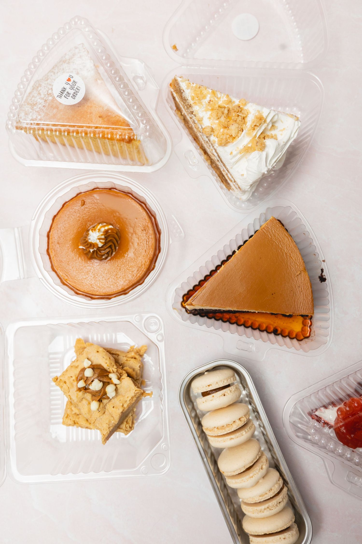 Assorted desserts in plastic containers including cheesecake slices, flan, macarons, and pastries on a white surface.