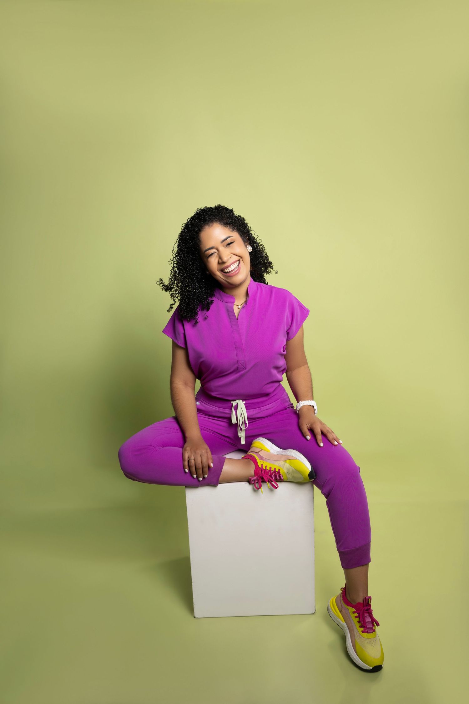 Person in bright purple athletic wear sits casually on white cube against lime green background.