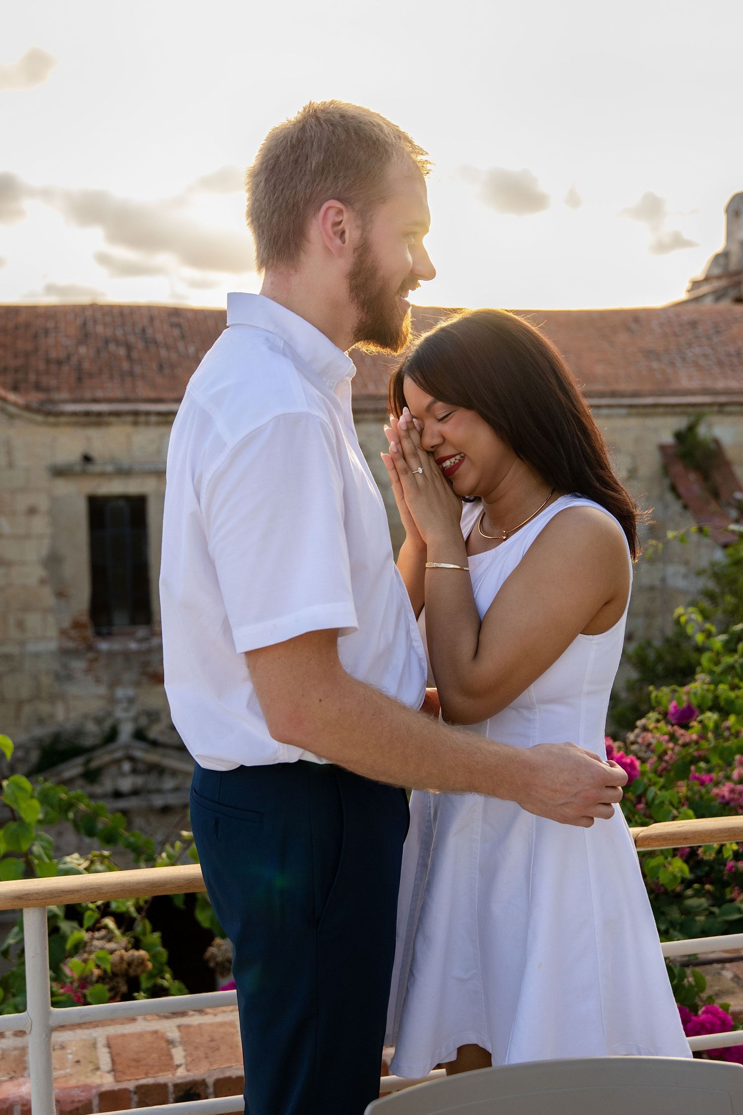 Couple in white outfits sharing a romantic moment on a balcony at sunset with historic building backdrop.